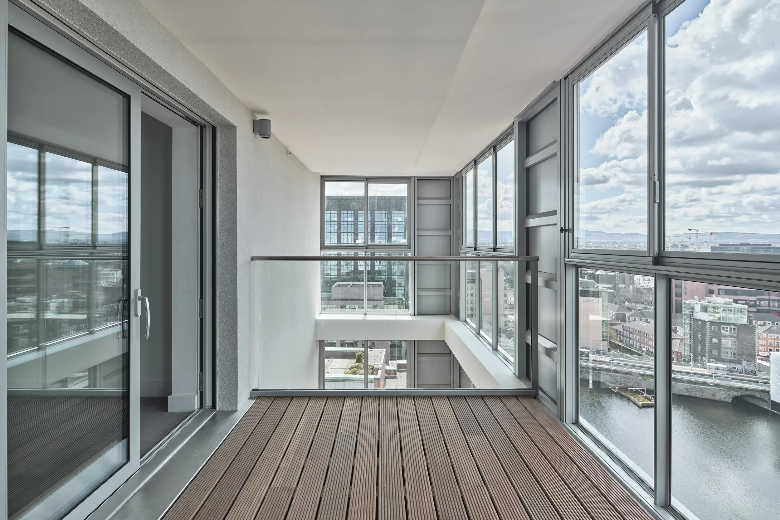 An empty balcony with wooden flooring, glass sliding doors, and large glass windows overlooking a cityscape with buildings under cloudy skies.
