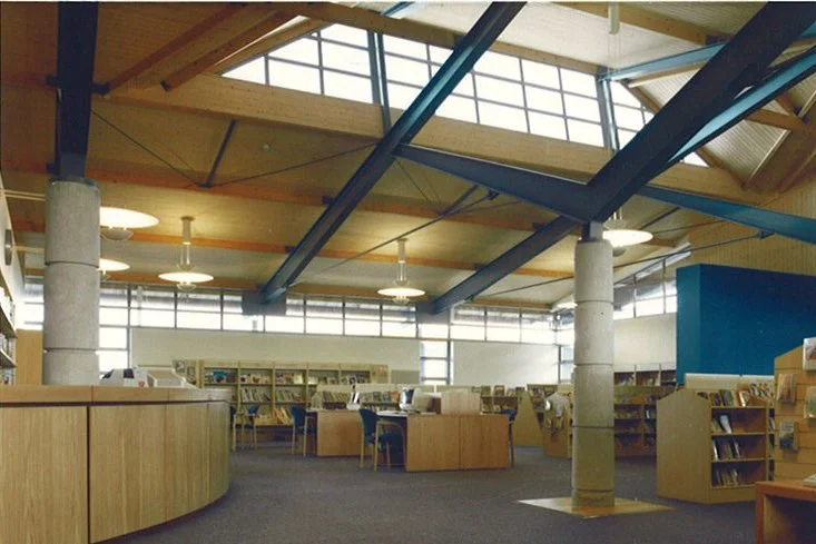 Interior of a modern library with wooden desks, bookshelves, large windows, and a high ceiling with exposed beams.