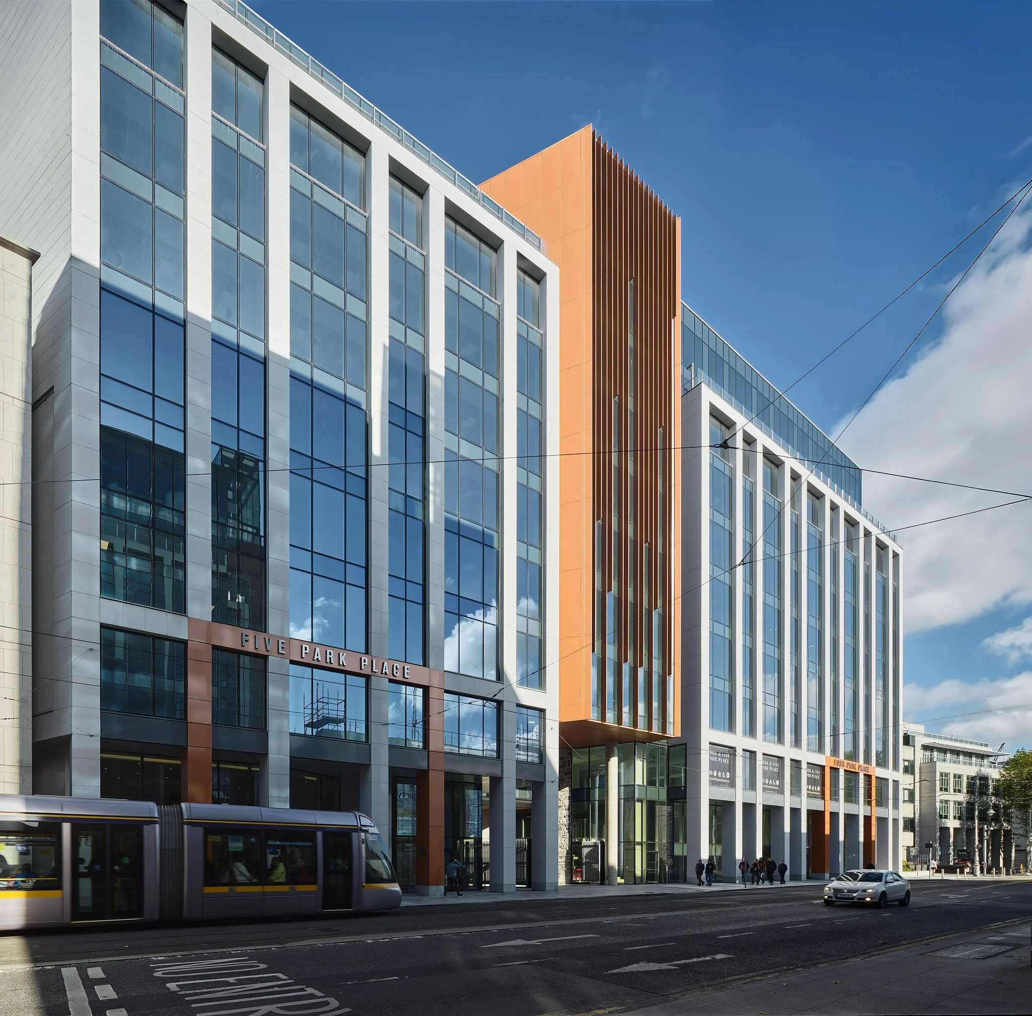 Modern multi-story building with glass windows and orange accents, sign reads 'Five Park Place', tram and cars on street, pedestrians walking along sidewalk, blue sky with clouds.