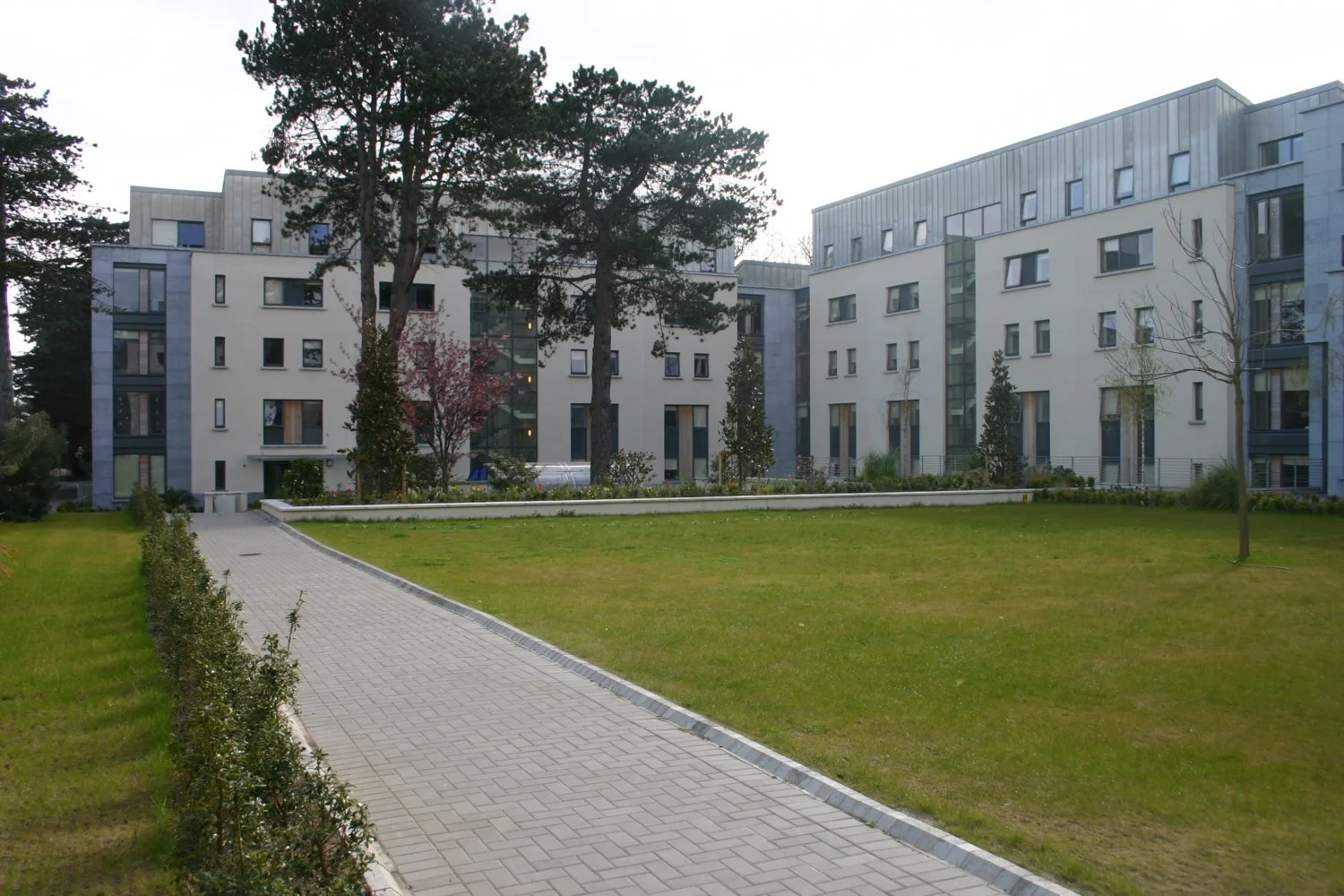 Modern apartment buildings with large windows, surrounded by well-maintained green lawns and trees, with a paved pathway leading through the garden.
