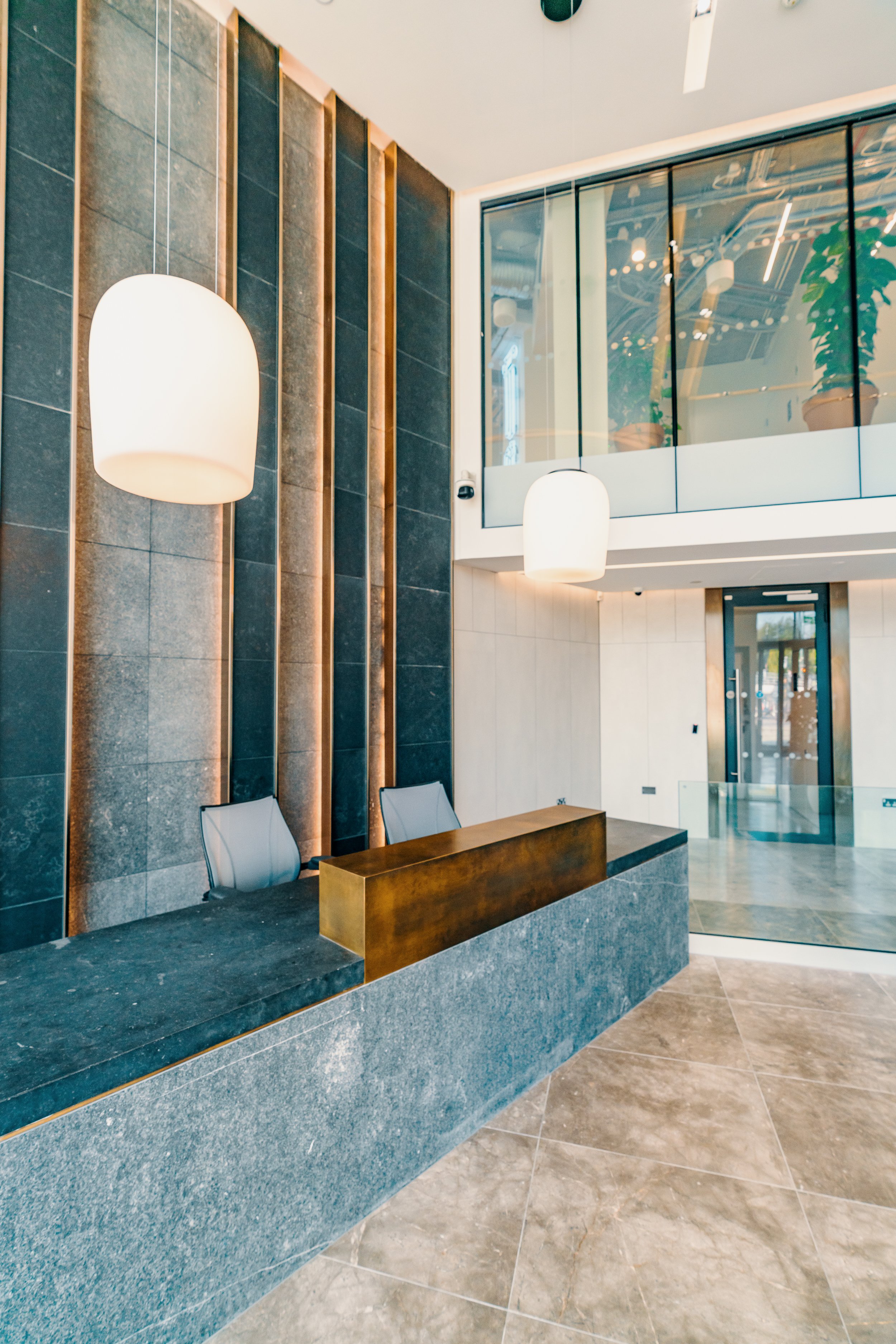 Modern hotel lobby reception area with a dark stone reception desk, two chairs behind it, hanging white pendant lights, a-glass balcony with plants, and a glass entrance door.