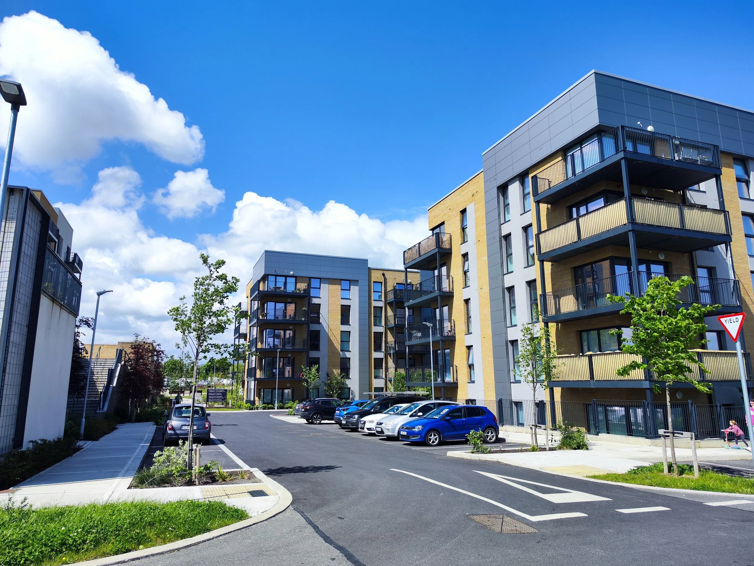Modern multi-story residential apartment buildings with parking lot and small trees under a blue sky with scattered clouds.