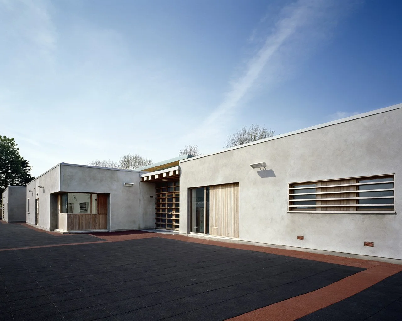 Modern white building with smooth exterior walls, large windows, wooden accents, and a paved area in front under a blue sky.