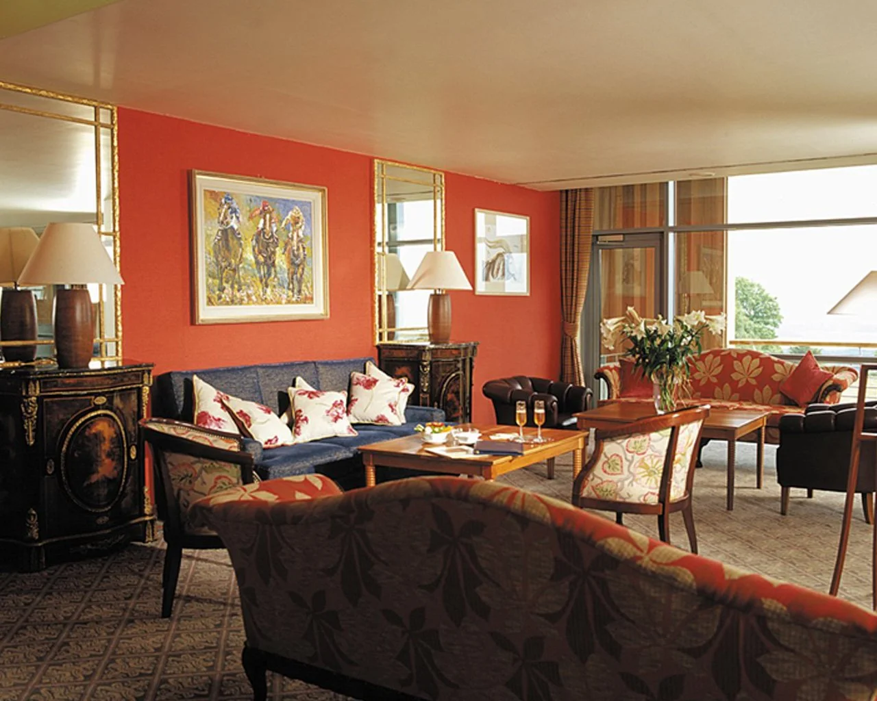 A luxurious and well-decorated living room with colorful furniture, artwork, and decorated with cushions, lamps, and a large window with curtains, a vase of flowers on a table.