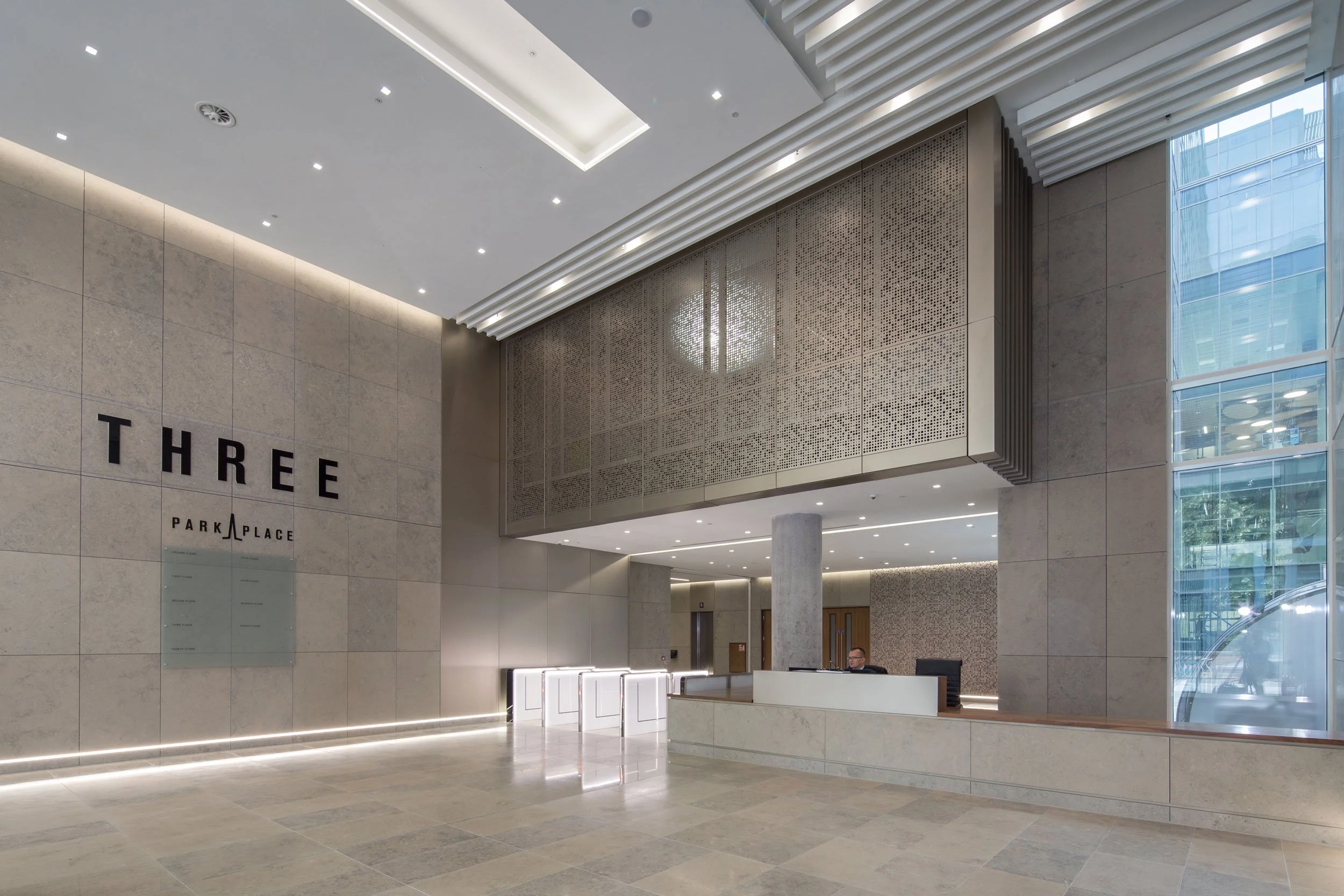 Modern hotel lobby with beige stone walls, a reception desk with a man, large glass windows, and contemporary lighting.