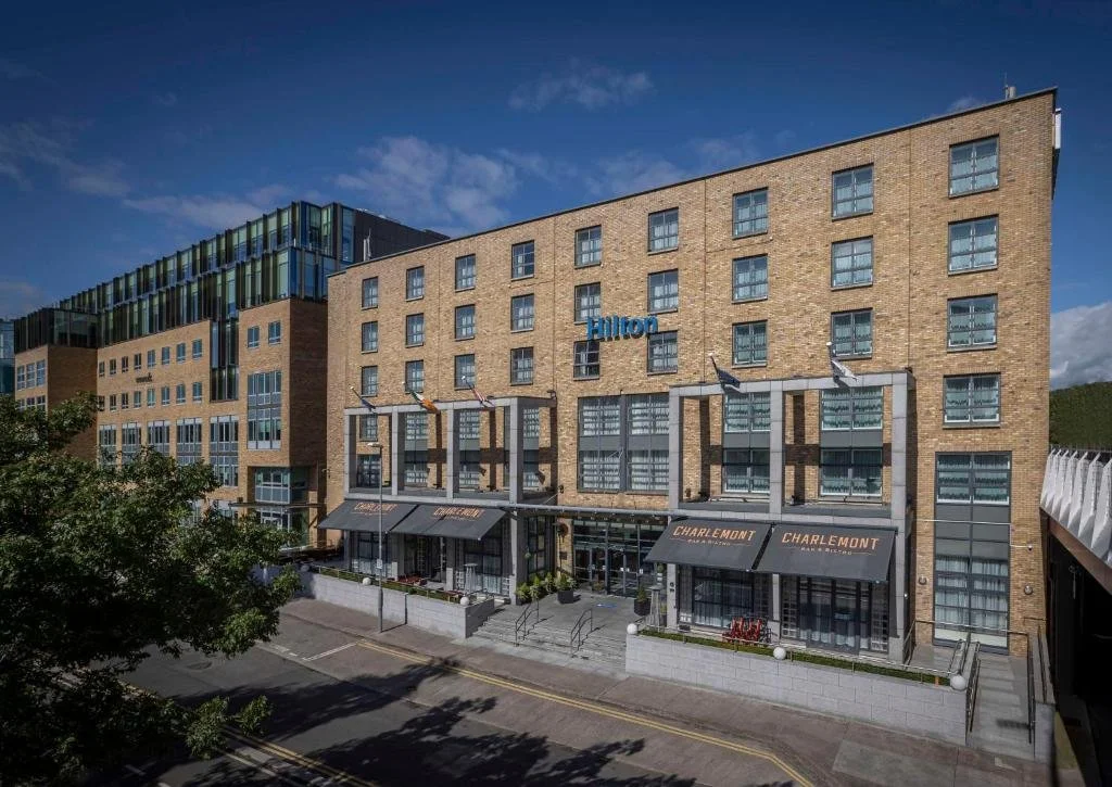Modern multi-story hotel with a brick facade, large windows, and outdoor seating area, located on a city street with a tree in the foreground.