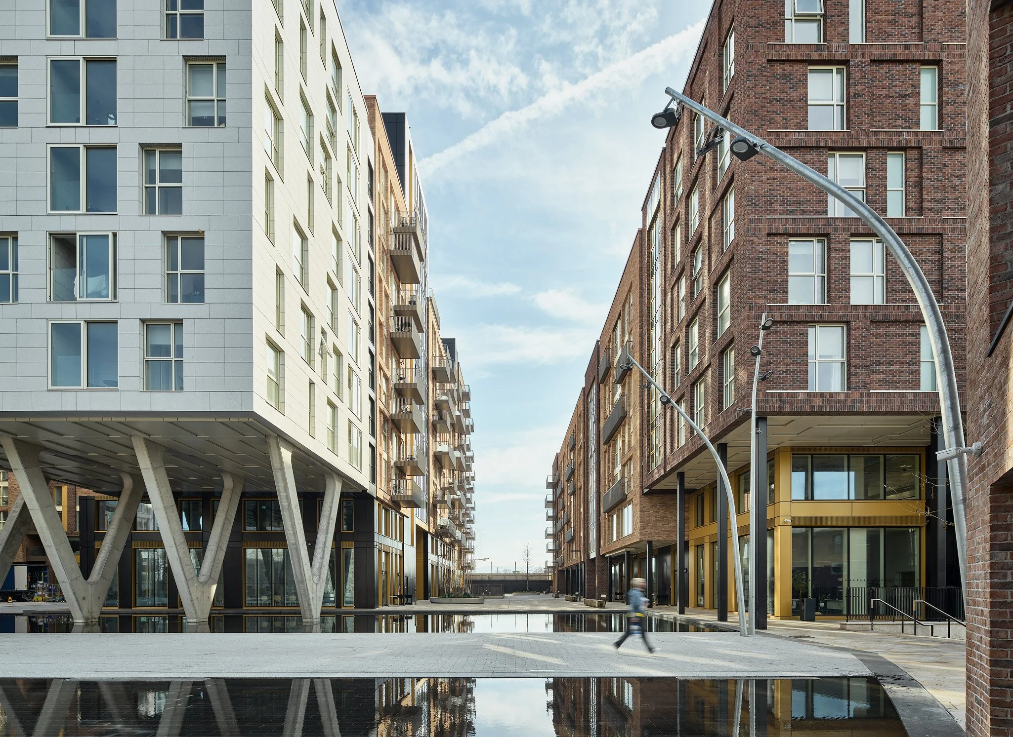 Modern urban street scene with tall buildings on either side, a person walking, and decorative street lamps, under a partly cloudy sky.