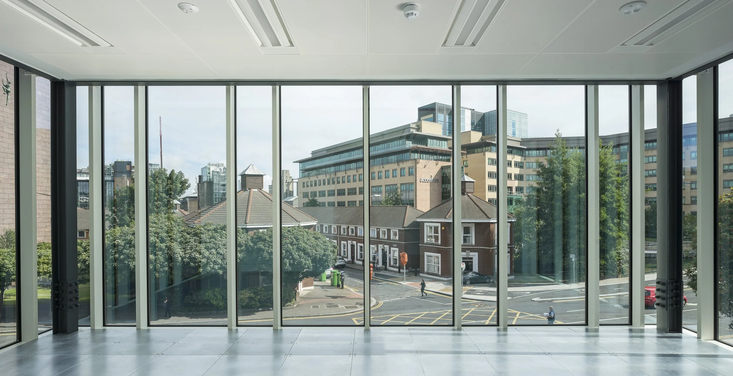 Interior view of a modern office with large glass windows, overlooking a city street with buildings, trees, and people walking.