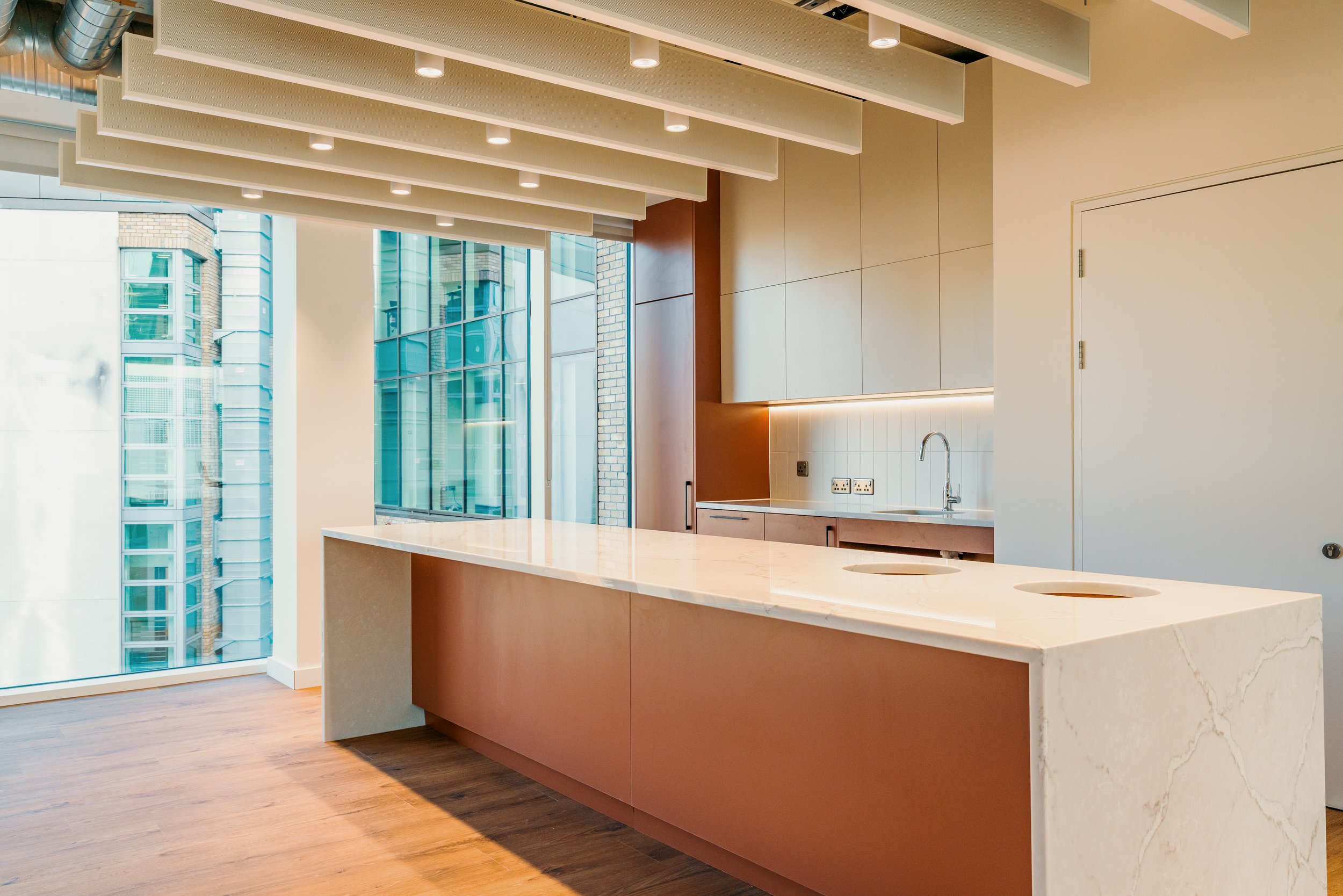 Modern kitchen with a large beige marble island, overhead lighting, and large windows showing an urban cityscape outside.