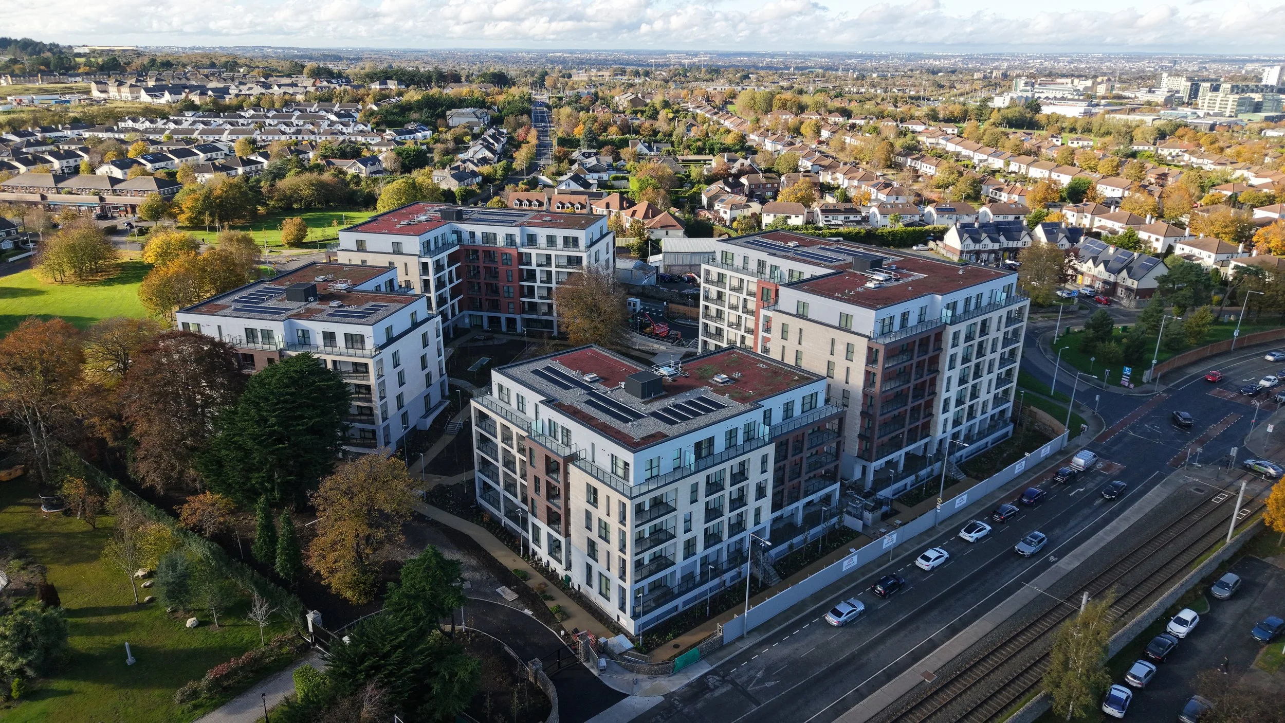 An aerial view of a residential area with modern apartment buildings, a parking lot, green spaces, and train tracks, during daytime with overcast sky.