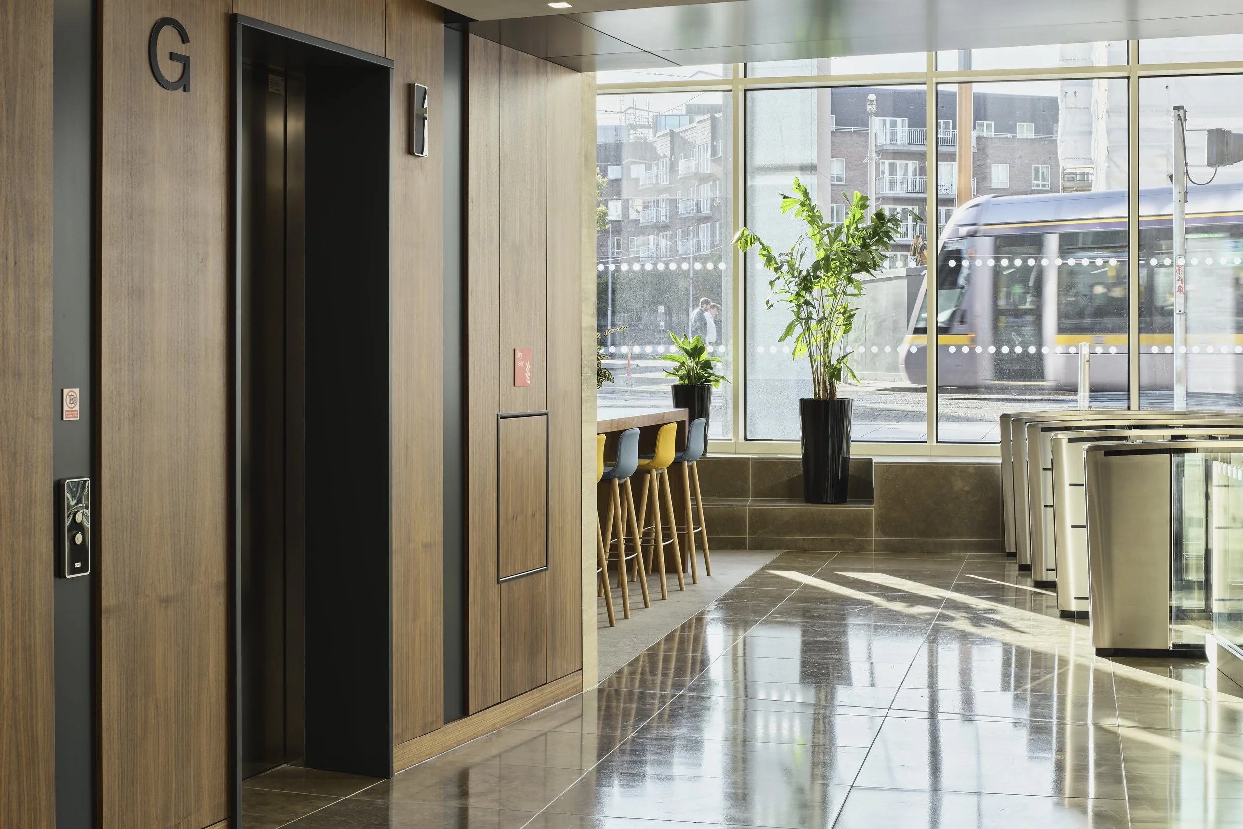 Interior of a modern building lobby with wooden paneling, an elevator, large windows letting in natural light, potted plants, bar stools, and a glass railing, with a city street and part of a tram outside.