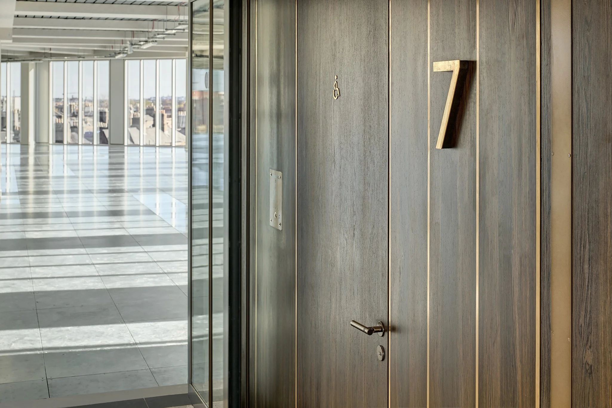 Modern elevator with wood paneling, a number 7 indicator, and accessibility symbols next to it in a high-rise building with a glass corridor.