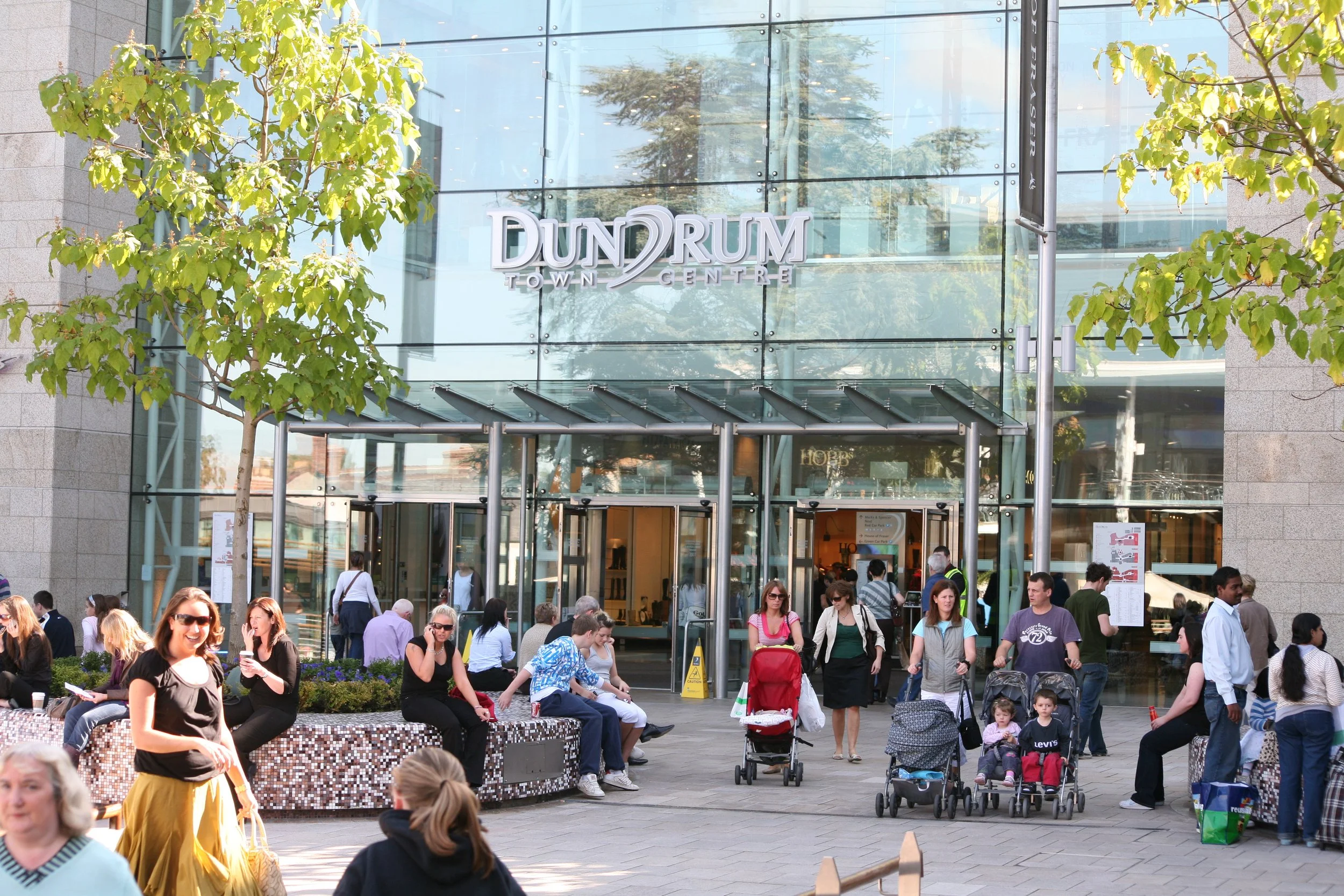 People gathered outside the Dunelm Town Centre store with some sitting and others pushing strollers in a busy shopping area with trees and glass building facade.