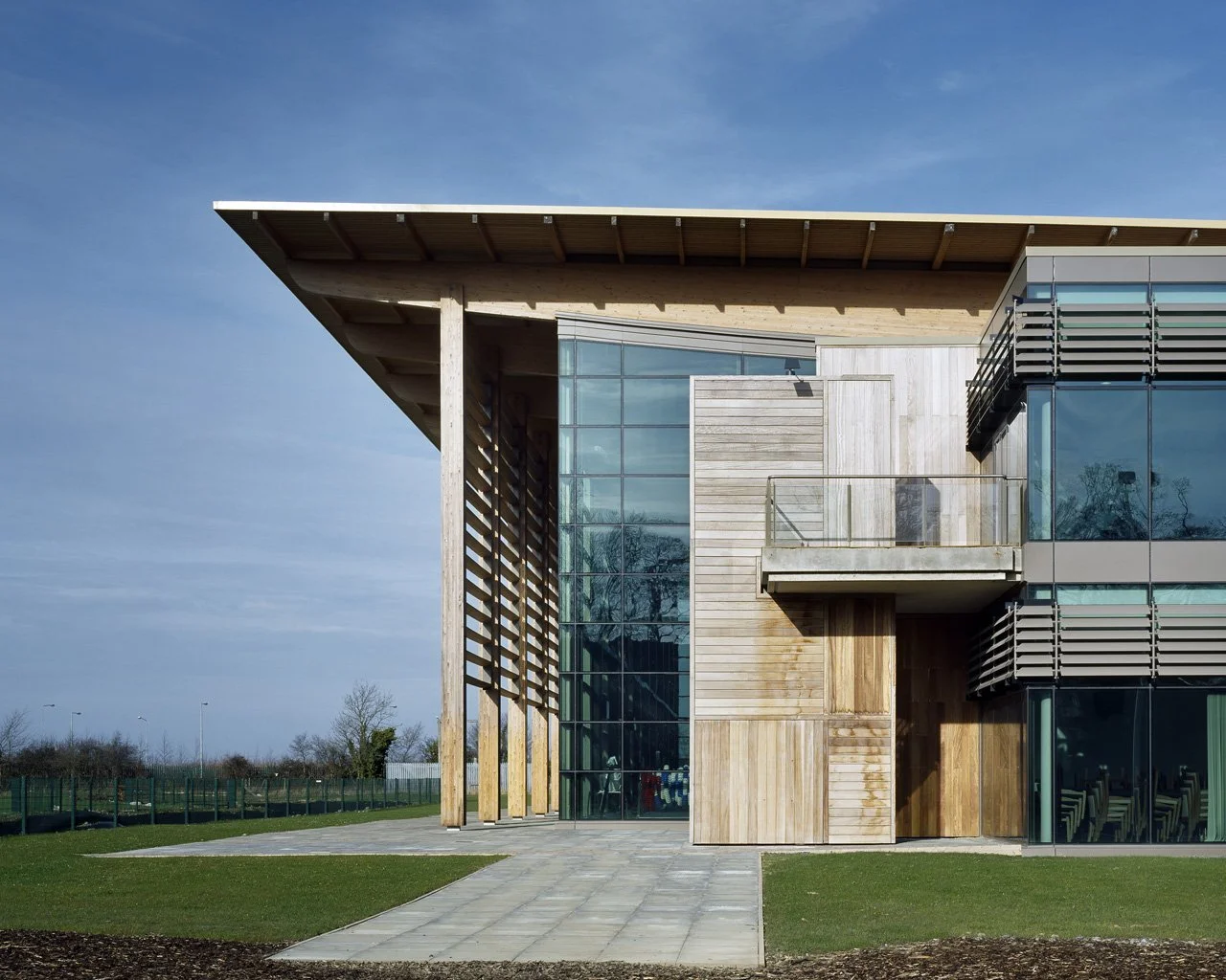 Modern building with a sloped roof, large glass windows, and wooden exterior panels, surrounded by grass and a paved walkway under a clear blue sky.