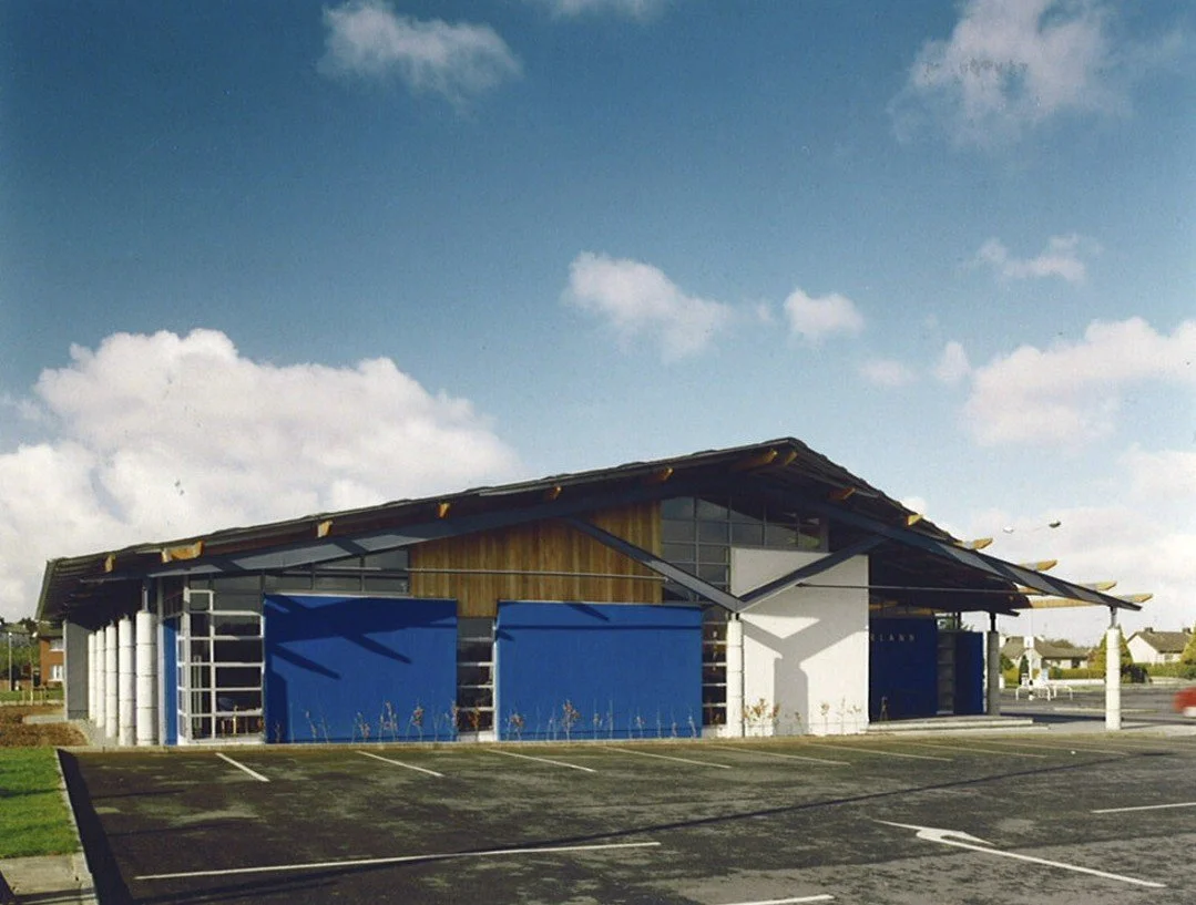 A modern building with a sloped roof, large windows, and blue and white exterior walls, next to a parking lot and a grassy area.