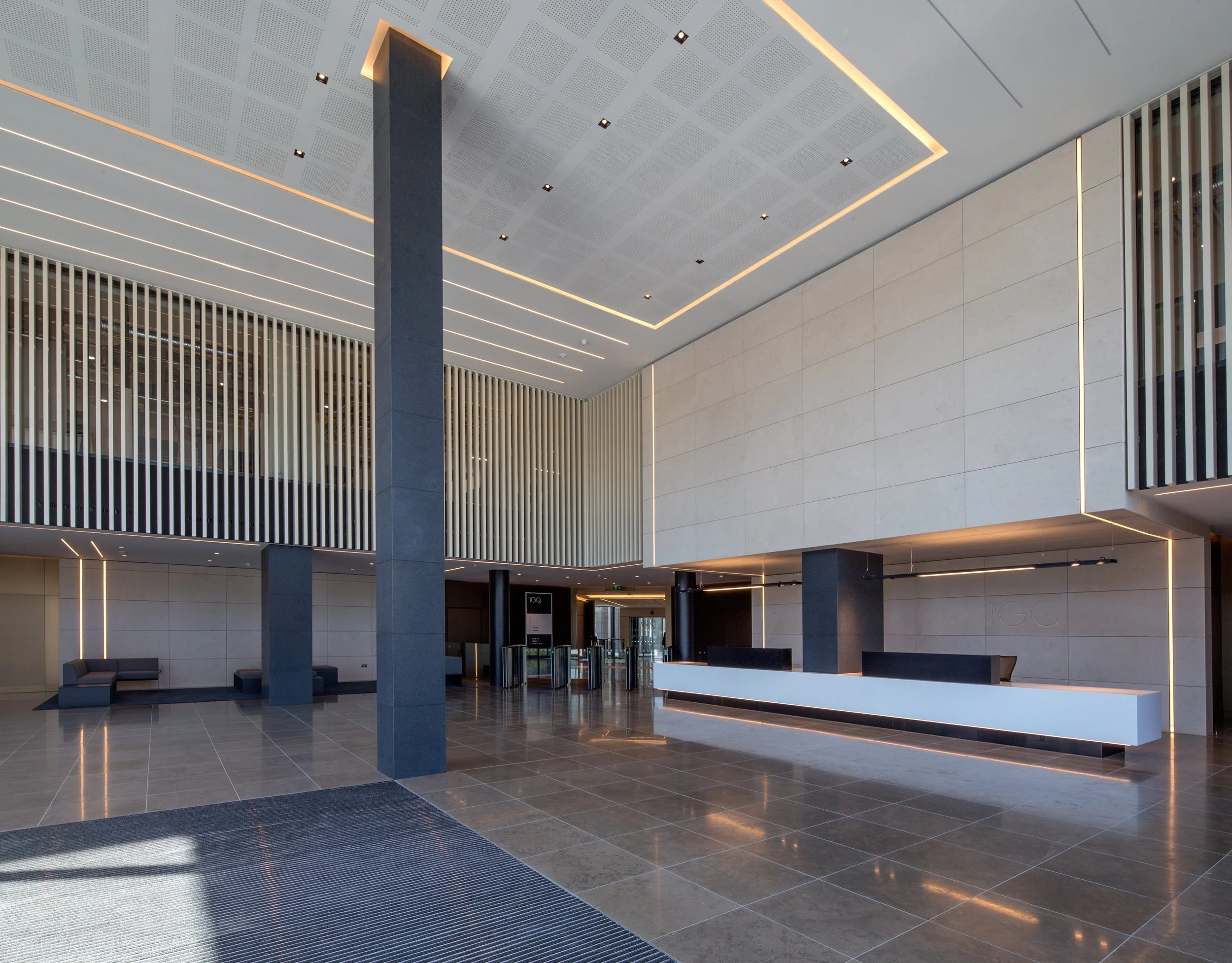 Modern hotel lobby with sleek black and white reception desk, seating area with black sofas, tall dark columns, and decorative vertical slats on the upper walls, illuminated with warm lighting.