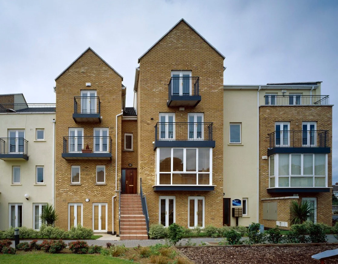 Multi-story apartment building with brick and cream-colored exteriors, balconies, and large windows, surrounded by small plants and a lawn, under a cloudy sky.
