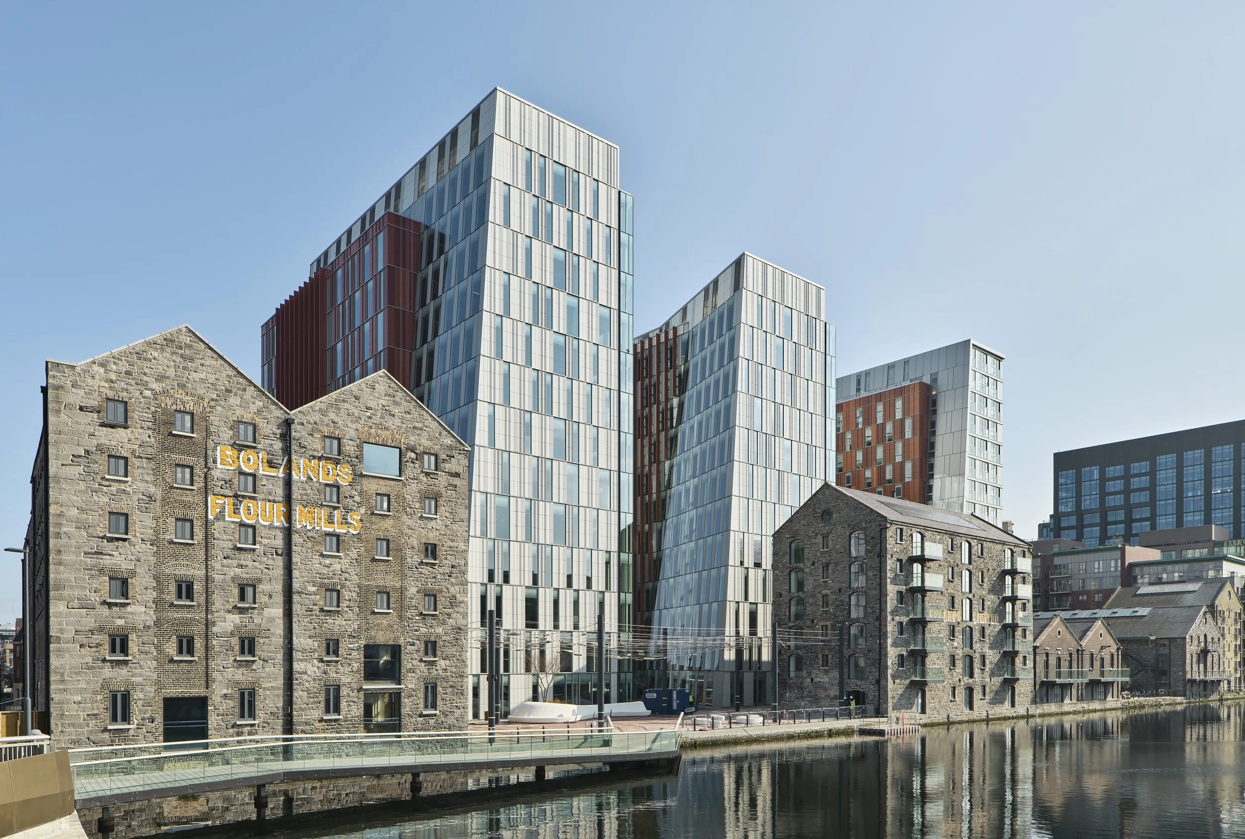 A modern cityscape with historic stone buildings along a canal, featuring a sign that reads 'BOLANDS FLOUR MILLS' and contemporary glass skyscrapers in the background under a clear blue sky.