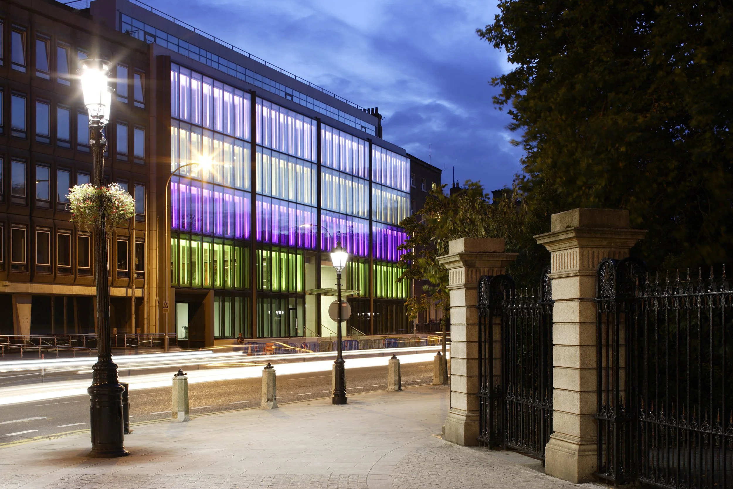 A modern building with colorful, illuminated glass panels at night, streetlights, a black iron fence, and light trails from passing vehicles.