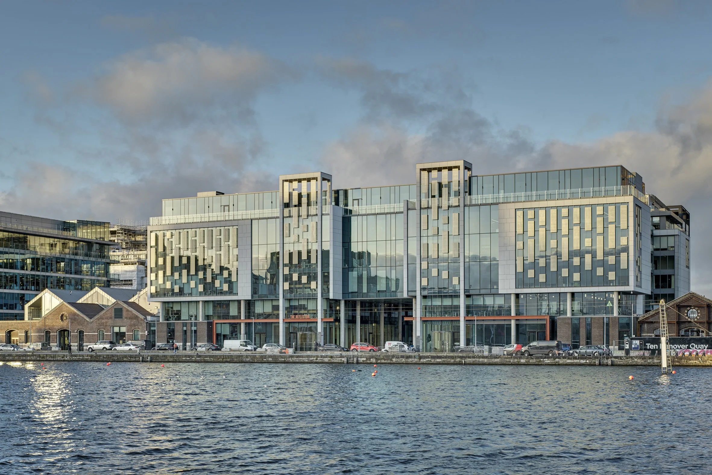 Modern glass building on water with boats and cars, partly cloudy sky.