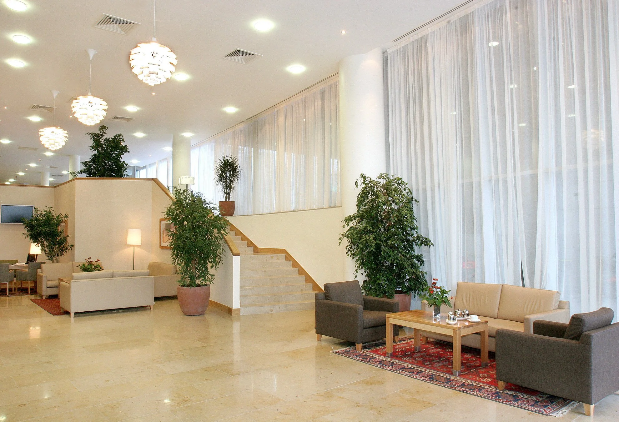 Hotel lobby with beige and brown furniture, potted plants, a staircase, and large windows with white curtains.