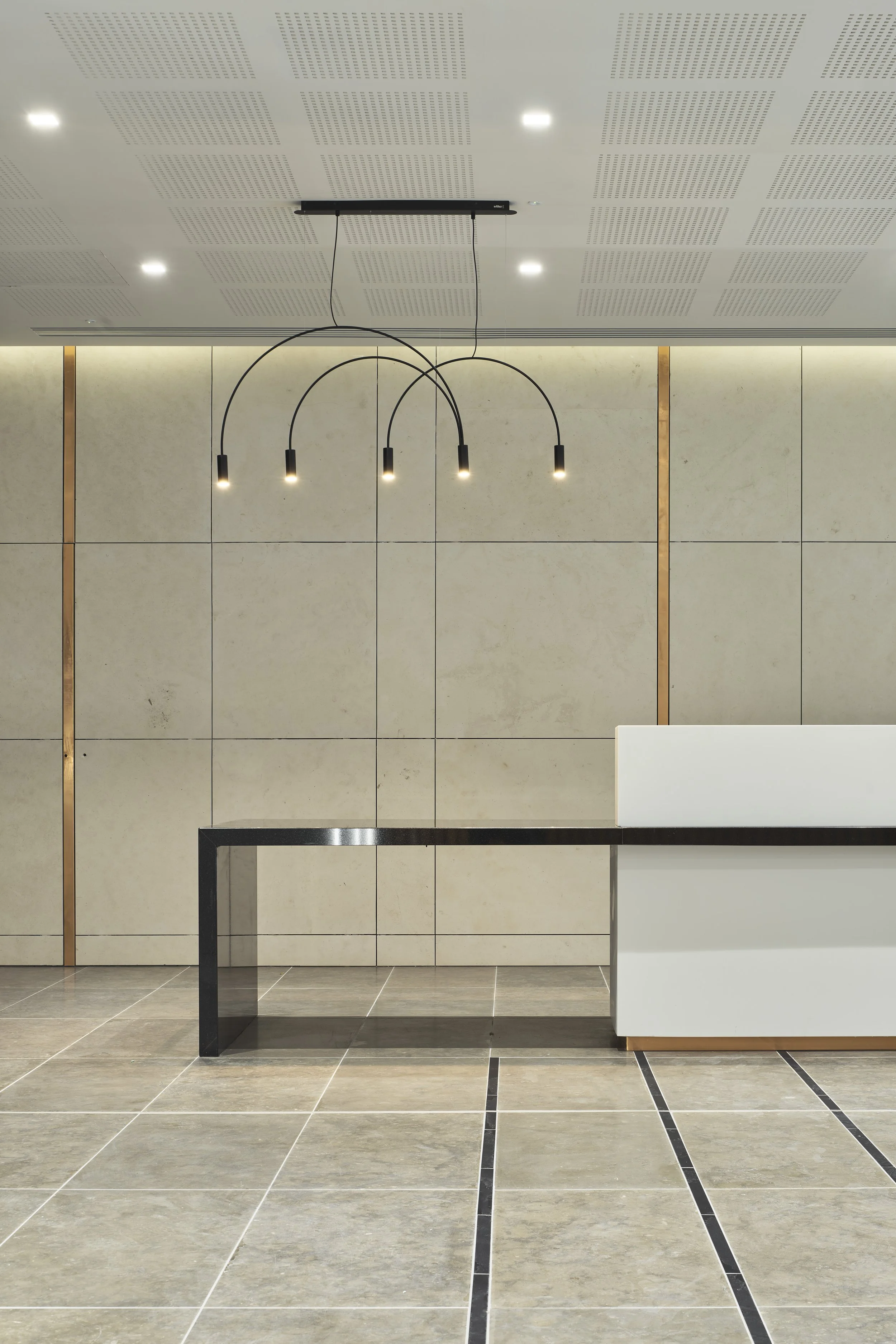 Modern lobby or reception area with beige tiled walls and floor, black pendant light fixture hanging from the ceiling, and a white reception desk with a black frame.
