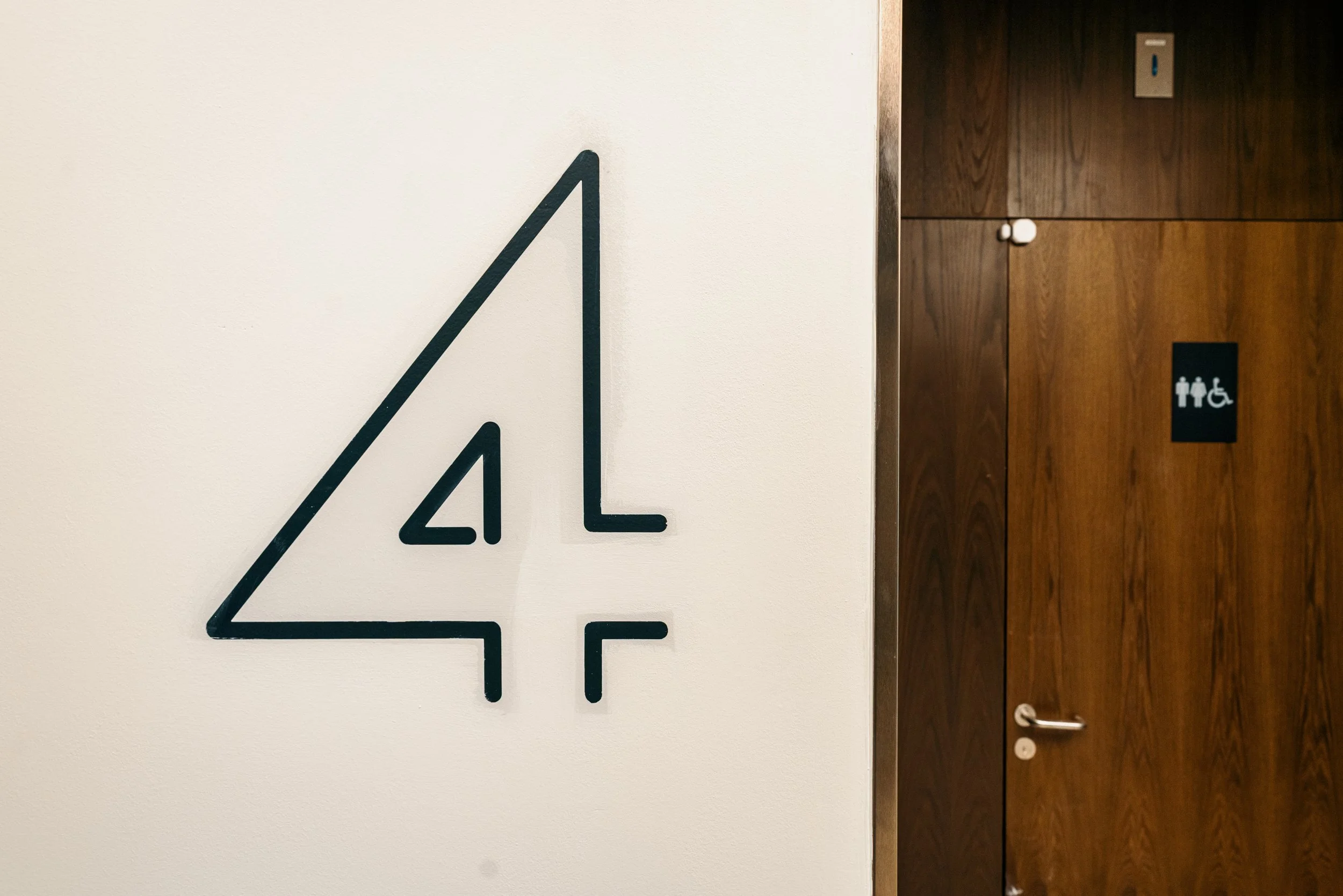 Sign indicating the fourth floor in a building, with a stylized number 4 enclosed in a triangle, next to a wooden door with a restroom sign.