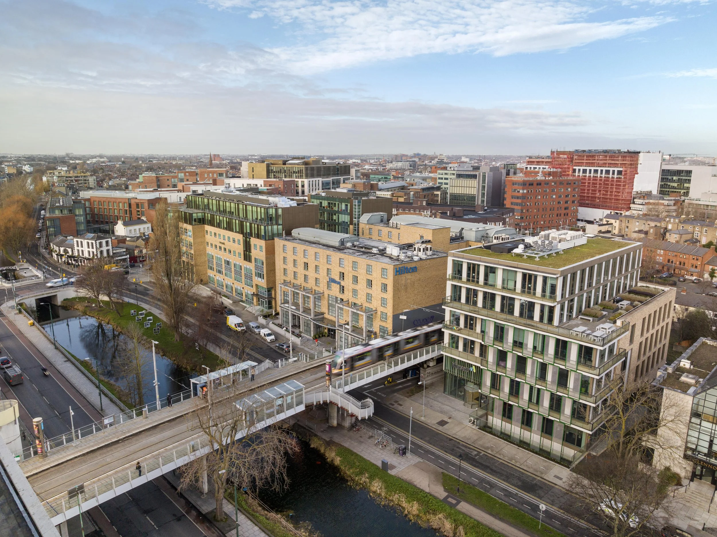 Cityscape with modern buildings, a rail bridge with a train passing, a canal, and streets in an urban area during daytime.