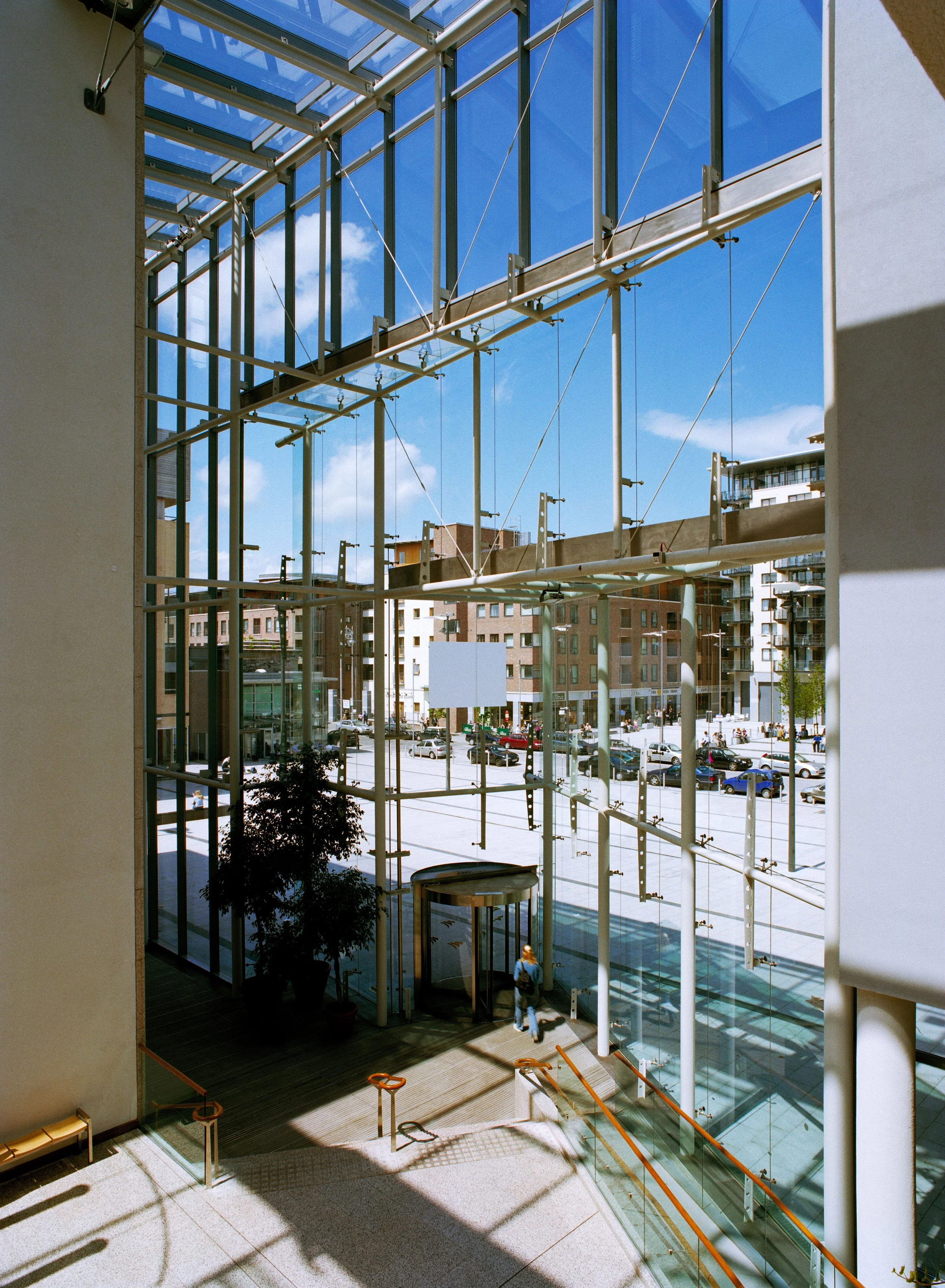 Photo taken from inside a building, showing a modern glass facade with steel framework, overlooking a city street with cars and apartment buildings.