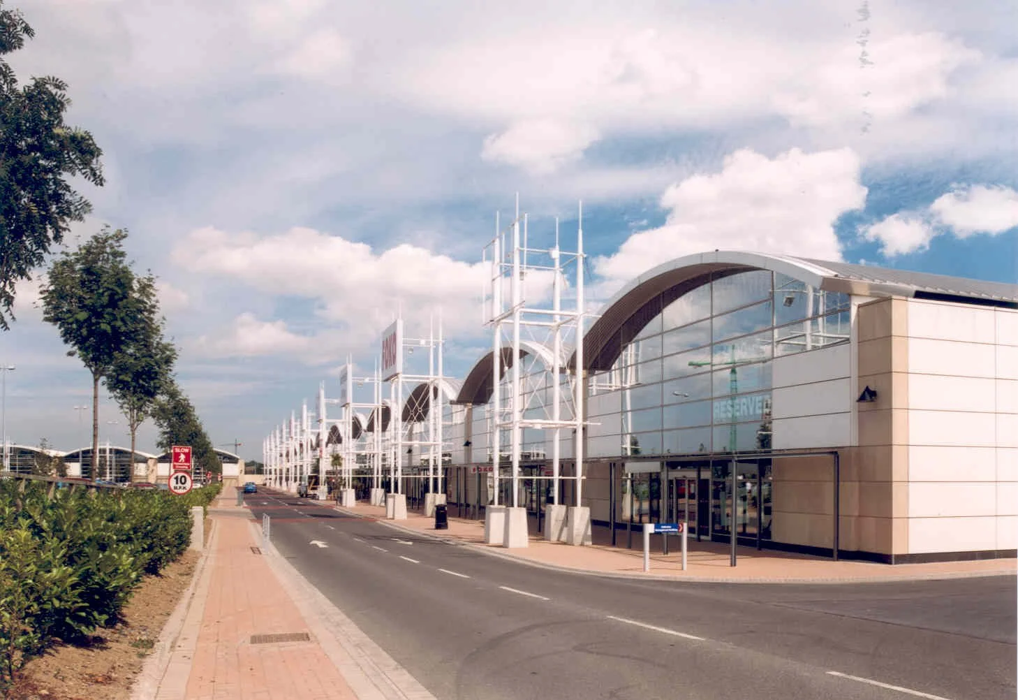 Modern shopping center with arched roof design, glass exterior, and pedestrian walkway, under a partly cloudy sky.