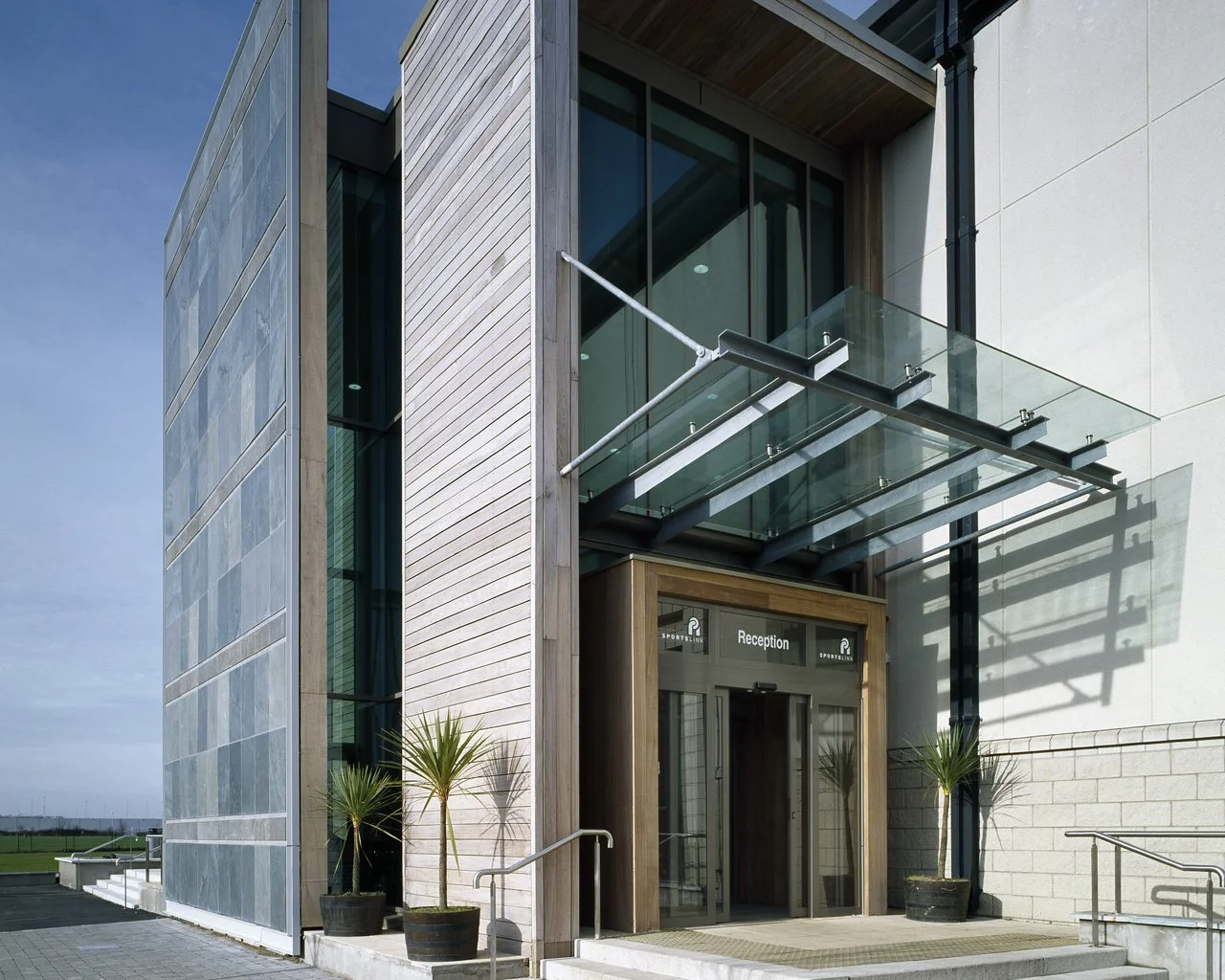 Modern building entrance with glass canopy, reception sign, potted plants, and stairs.