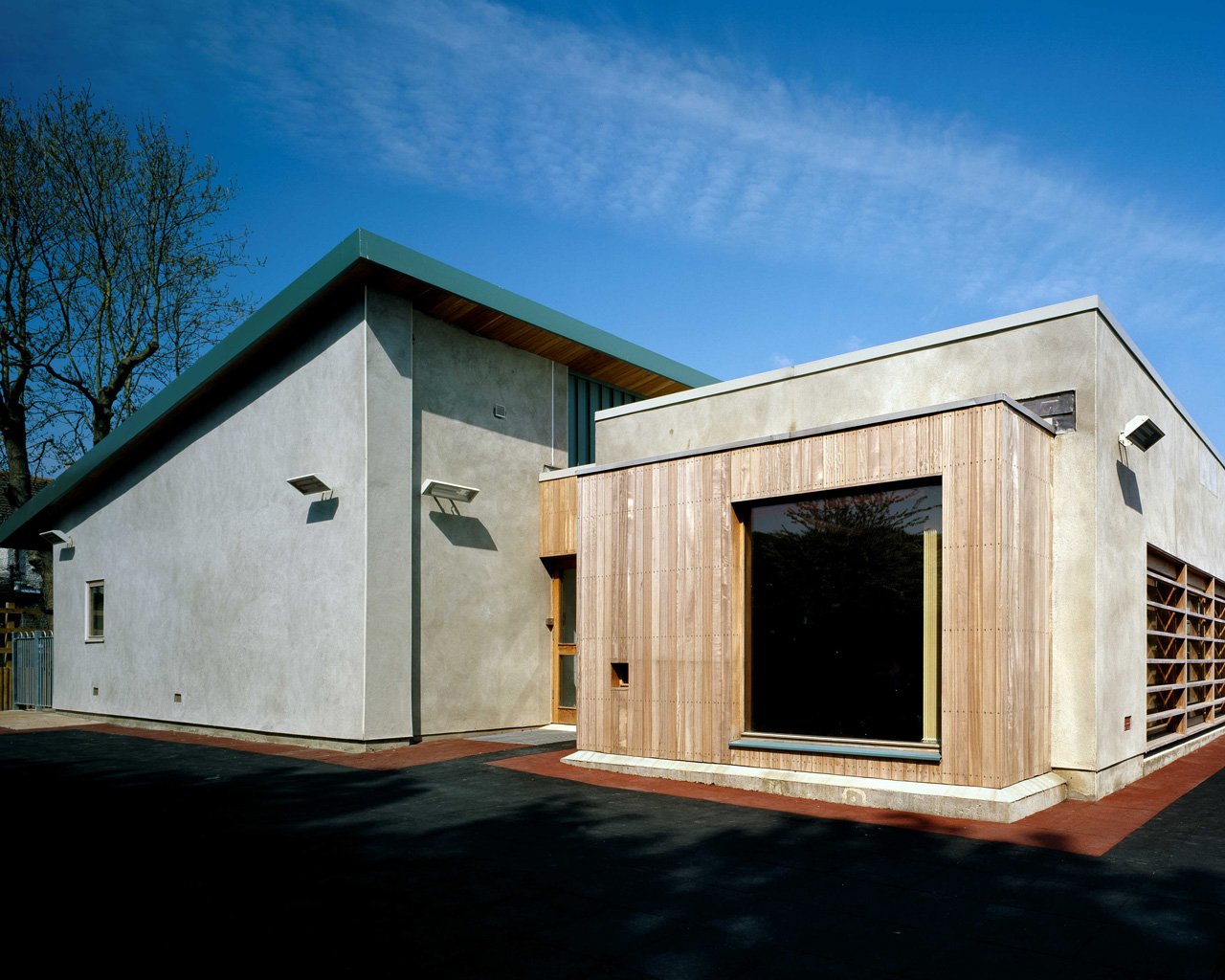 Modern two-story house with concrete and wooden exterior, large window, and sloped green roof, under a blue sky.