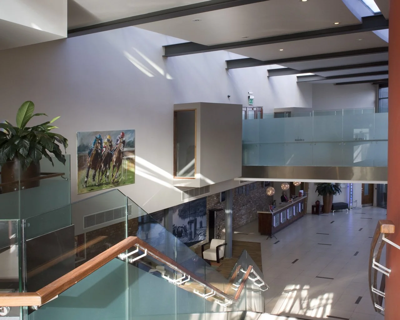 Interior view of a modern building with a staircase, large potted plants, a horse race painting, and a reception desk, illuminated by natural light from skylights.