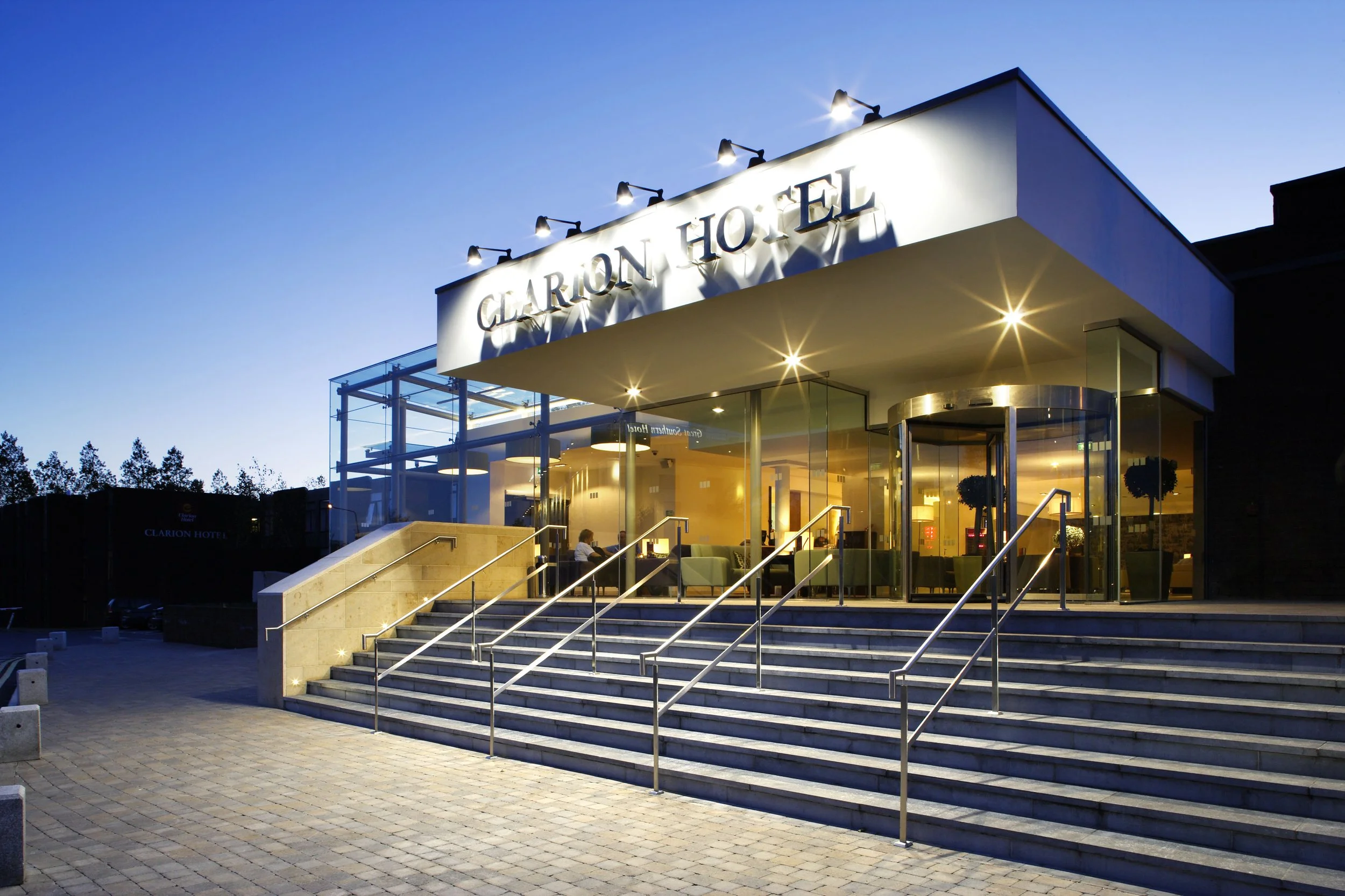 Exterior of the Clarion Hotel with glass doors and wide staircase, evening scene with illuminated sign and lights inside.