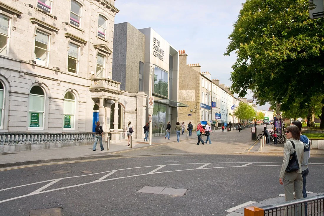People walking on a city street with shops and trees, some buildings with signs, and a large modern building labeled 'EYE SQUARE CENTRE'.