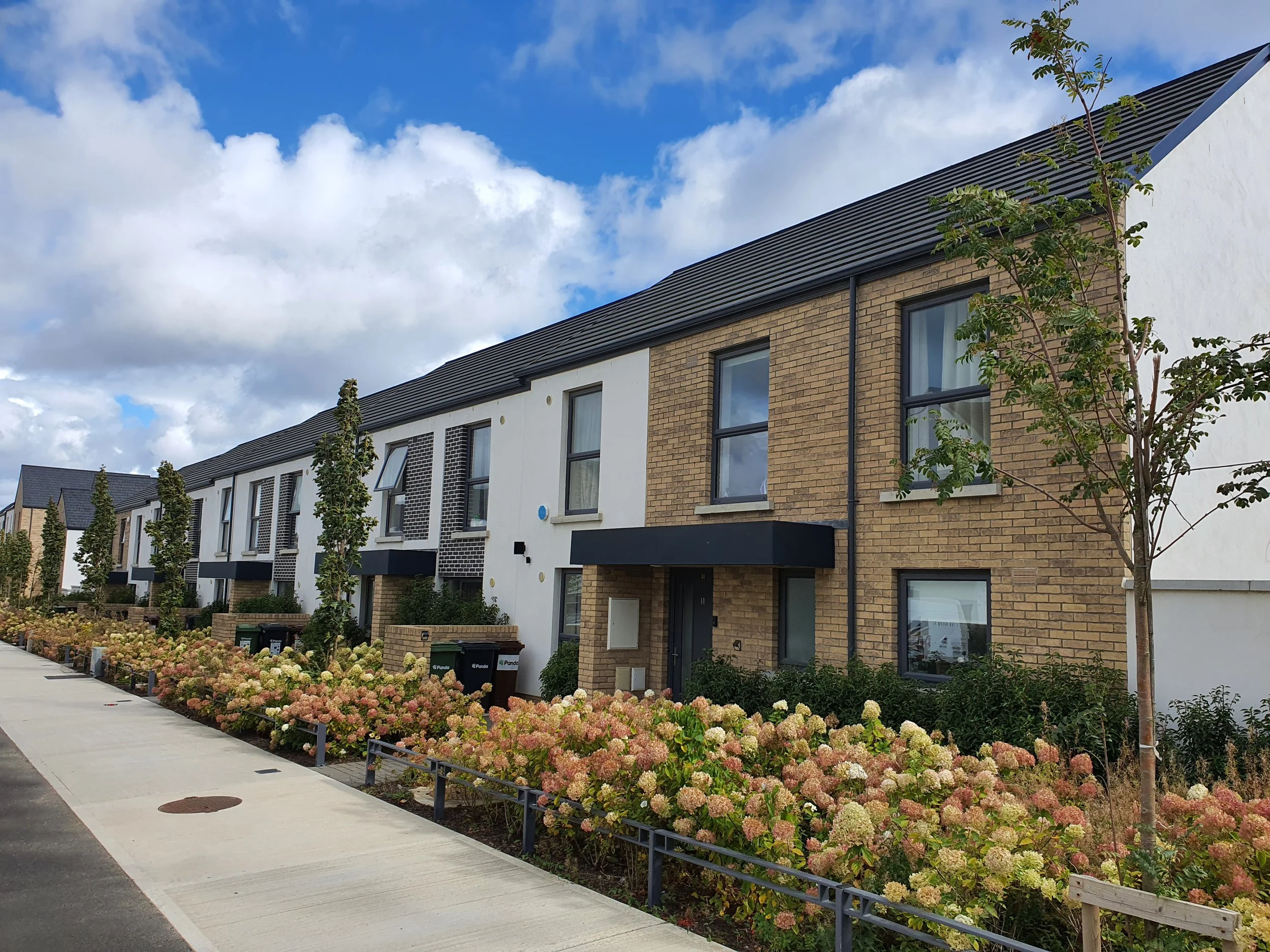 Modern row of residential townhouses with brick and white facades, trees, colorful flowering bushes, and a sidewalk in the foreground under a partly cloudy sky.