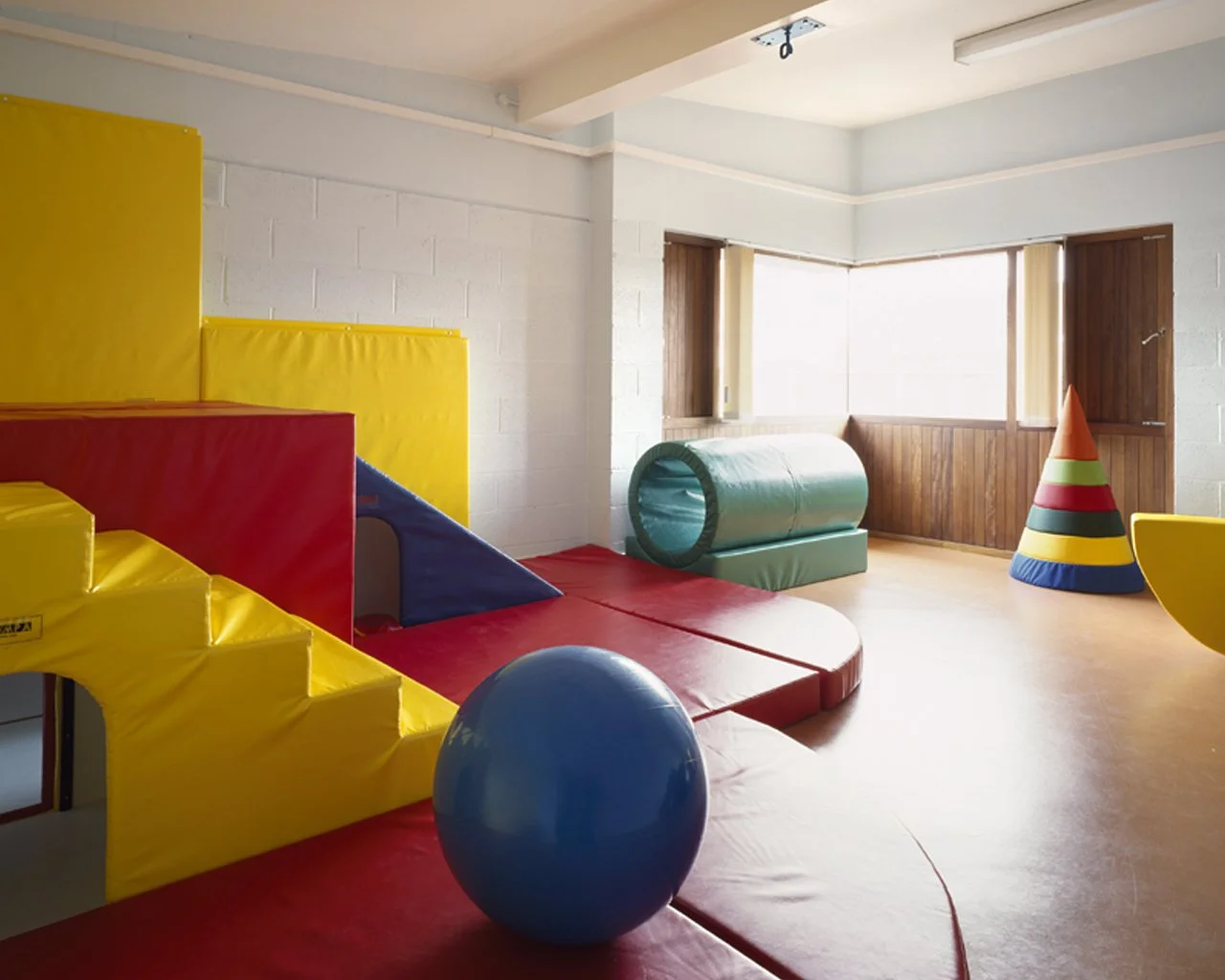 Children's indoor playroom with colorful foam climbing structures, a large blue ball, a green tunnel, and a cone-shaped soft play item in the corner.