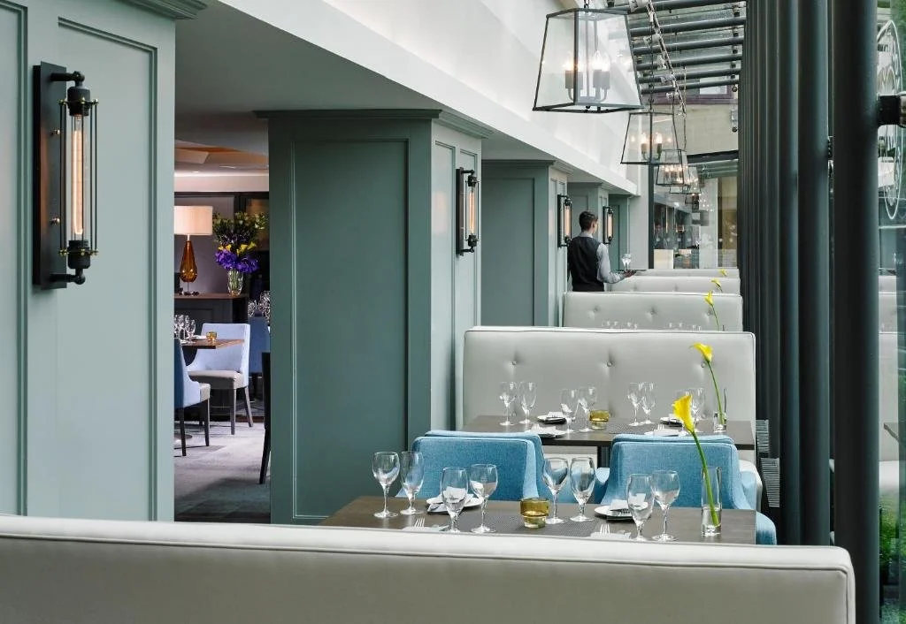 Upscale restaurant interior with white and blue booths, glassware on tables, and modern light fixtures. A server is seen in the background.