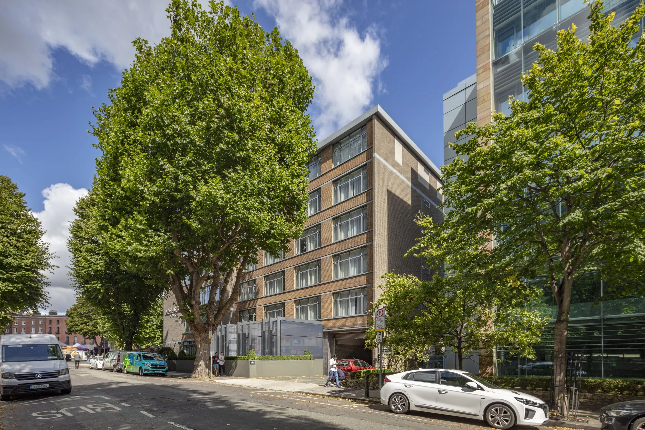 Street view with parked cars, large trees, and a multi-story brick building with a glass entrance on a sunny day.