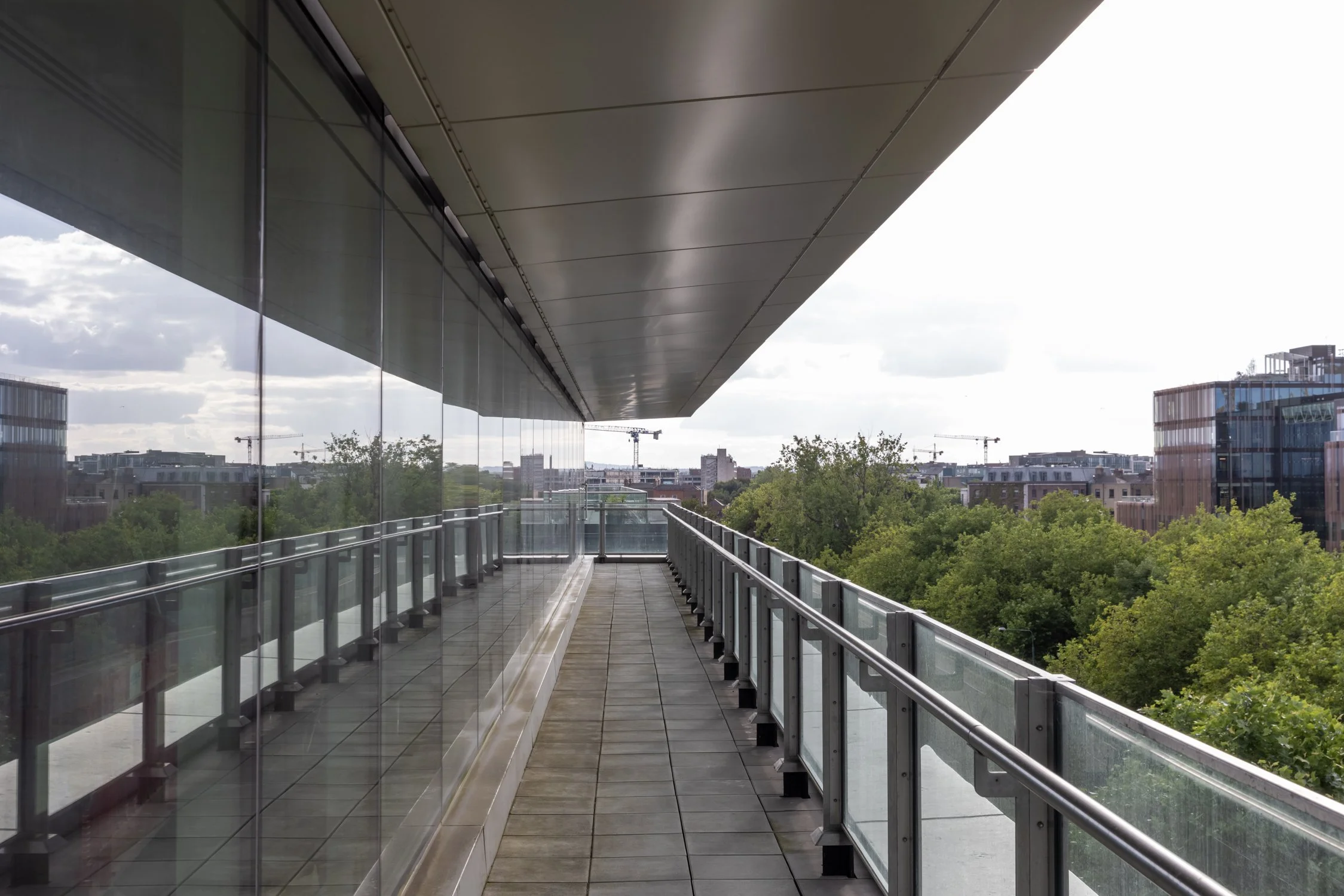 Empty outdoor balcony with glass railing, overlooking city buildings and trees, under a cloudy sky.