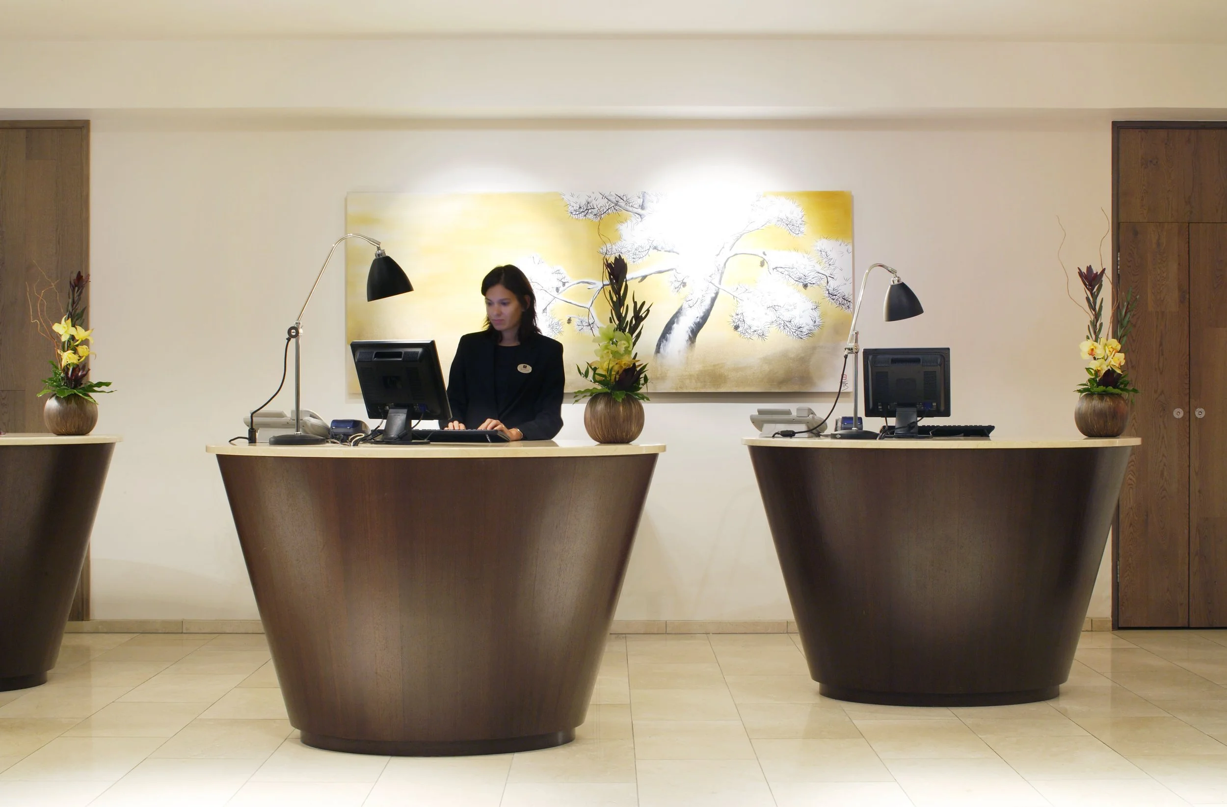 Hotel reception desk with a woman receptionist working at her computer, flanked by two vases with flowers, and a large artwork of a tree on the wall behind her.