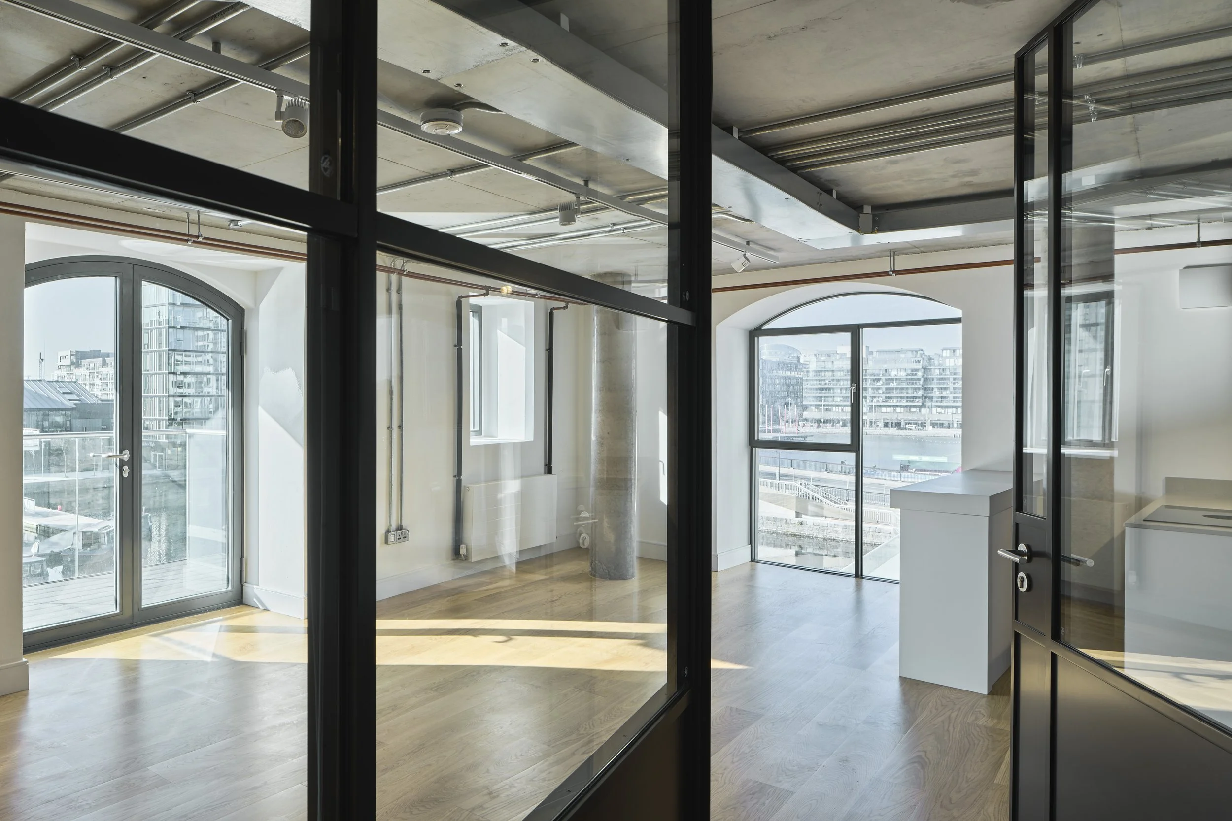Empty modern apartment with large windows, wooden floors, and exposed ceiling with pipes and conduit, viewed through glass doors.
