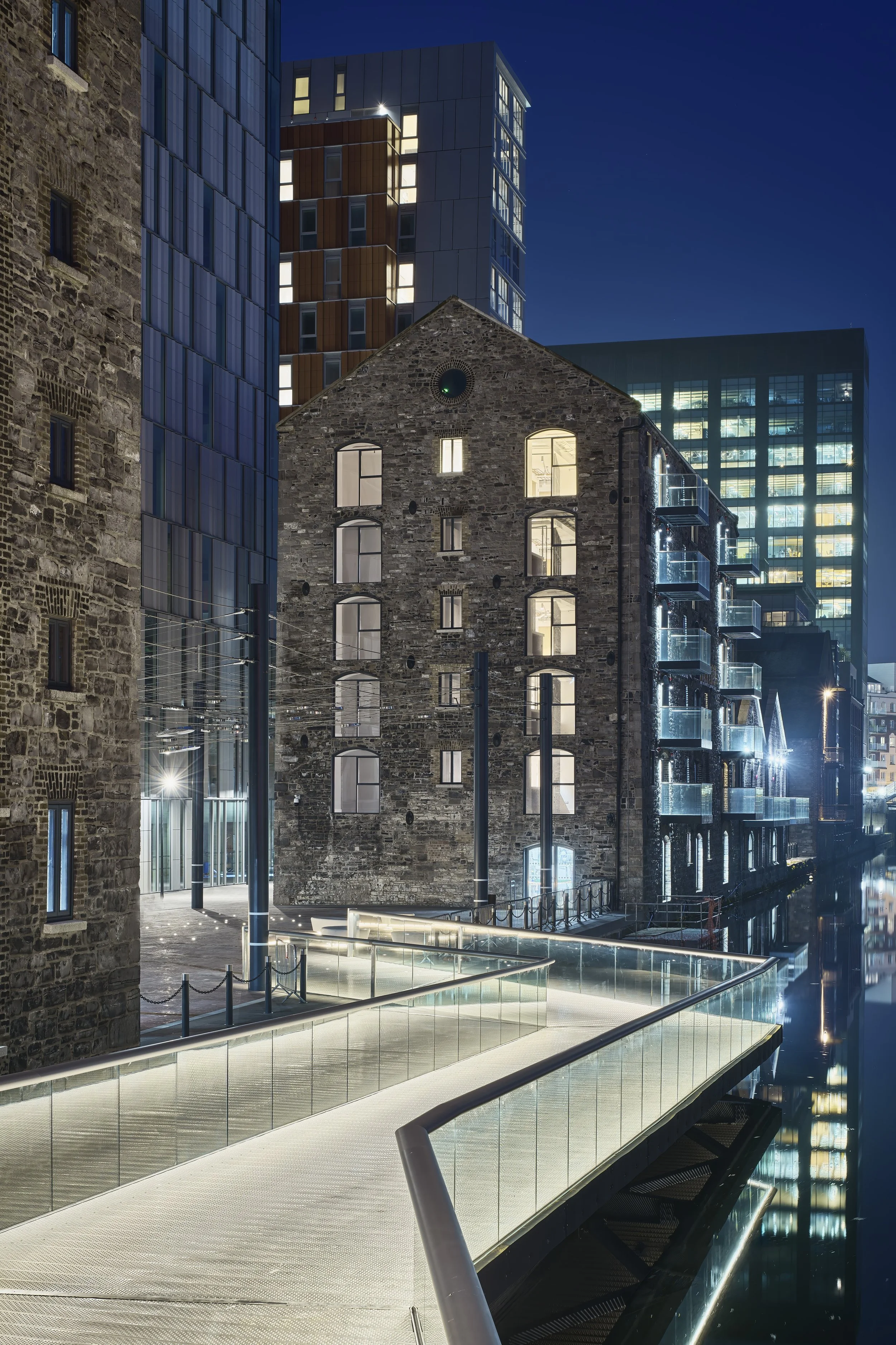 Nighttime cityscape featuring a modern glass walkway leading to a historic brick building surrounded by tall contemporary office buildings with illuminated windows.