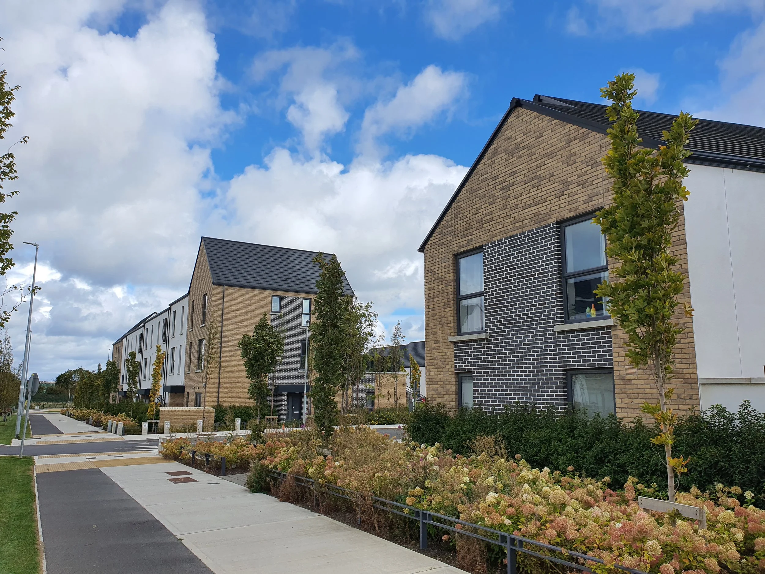 New modern residential neighborhood with townhouses, trees, and a sidewalk under a partly cloudy sky.