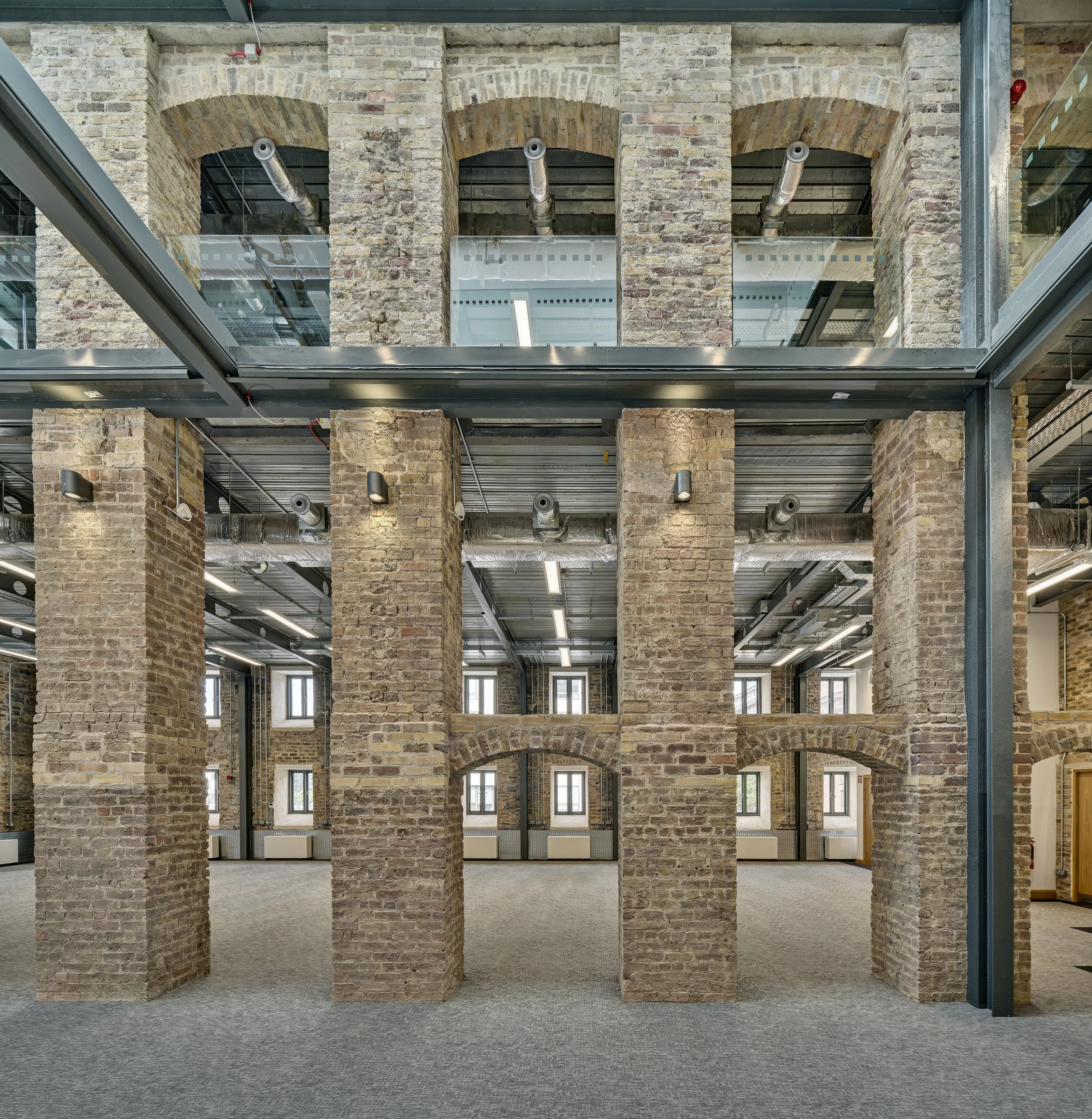 Interior of a brick building with exposed brick walls, large brick columns, and visible industrial piping and ductwork on the ceiling. Multiple windows with natural light, and a carpeted floor.