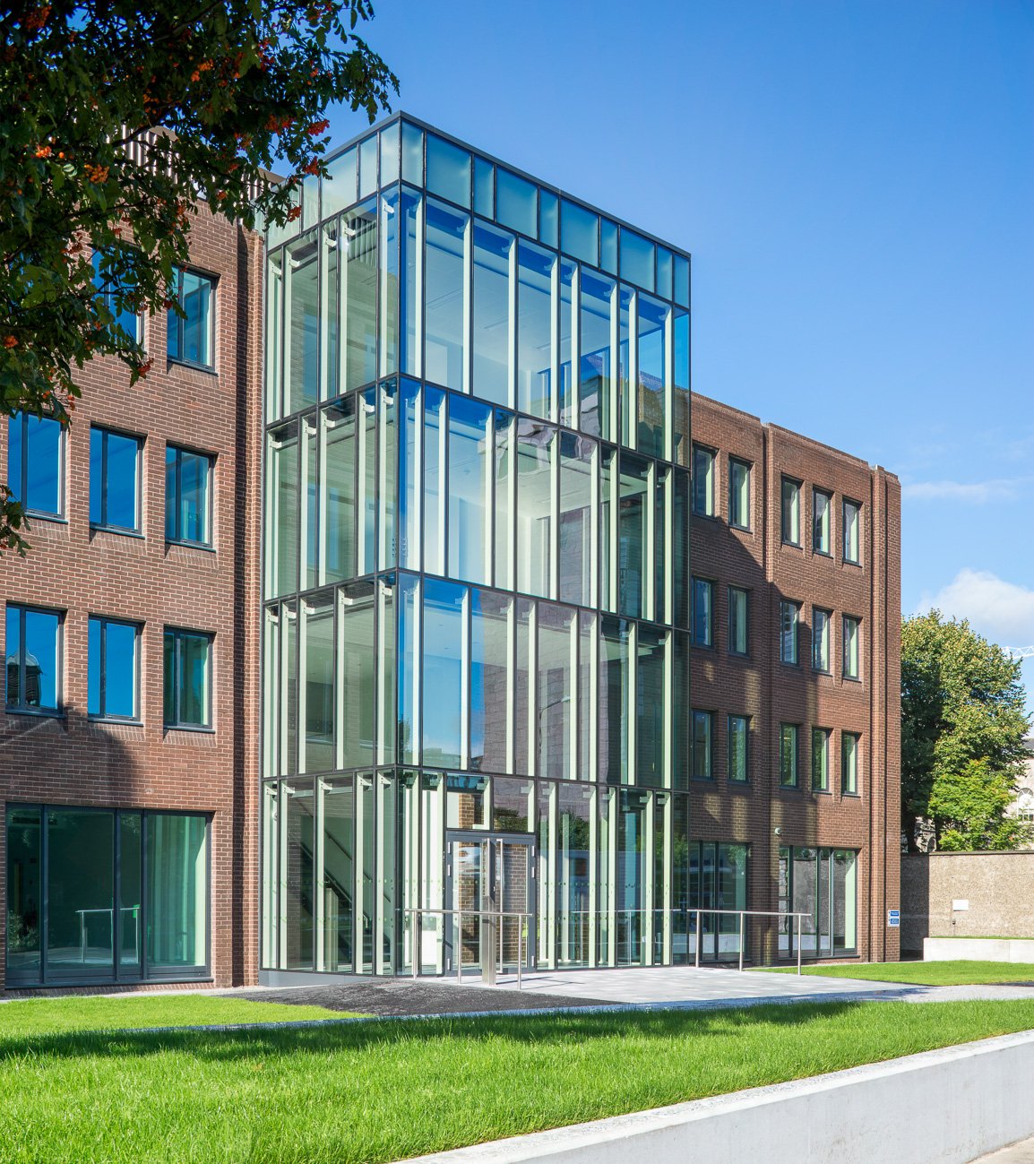 Modern brick building with glass staircase extension and lush green lawn under a clear blue sky.