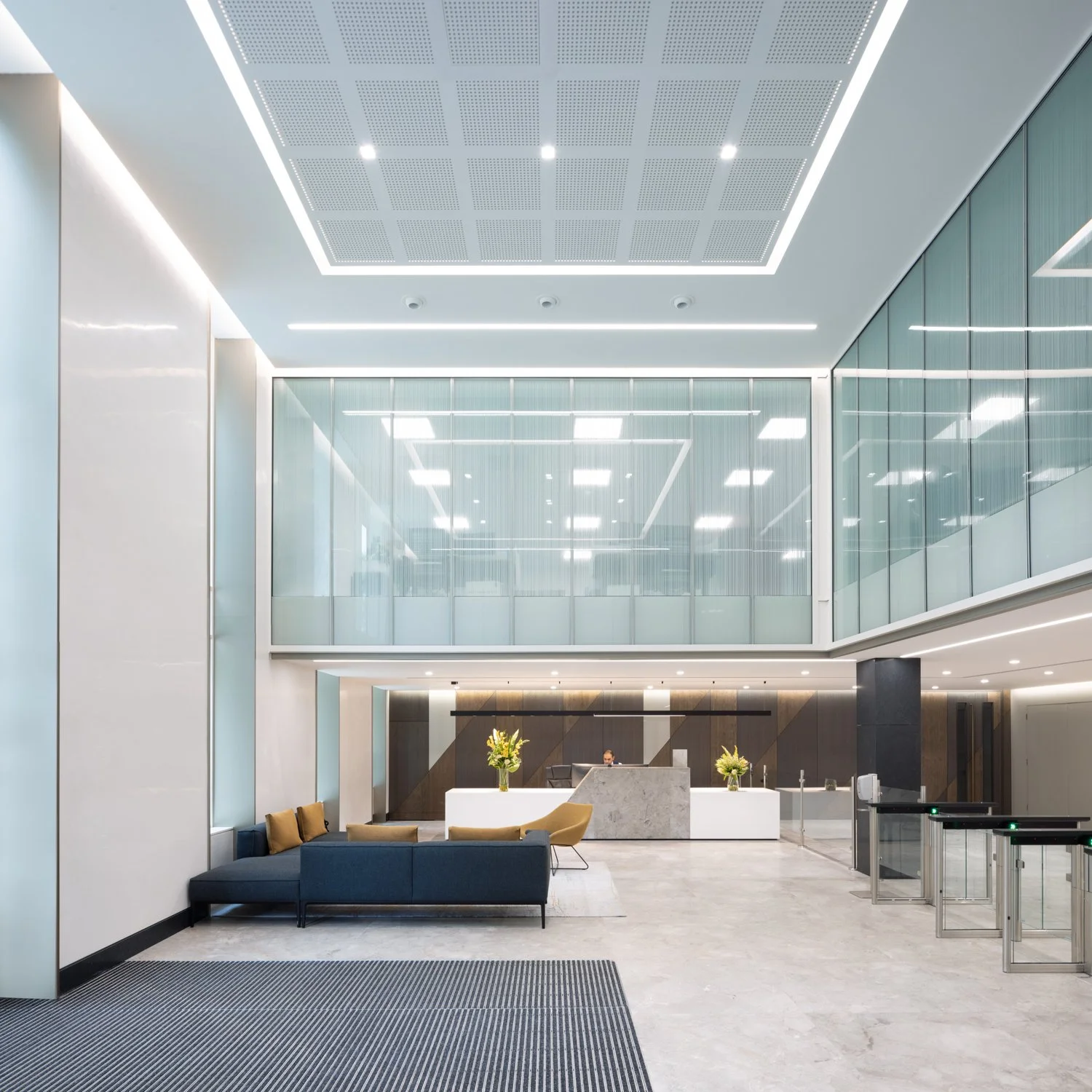 Modern office lobby with seating area, reception desk, glass walls, and ceiling lighting.