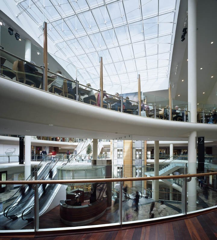 Multiple floors of a modern shopping mall with glass railings, escalators, and people walking and shopping.