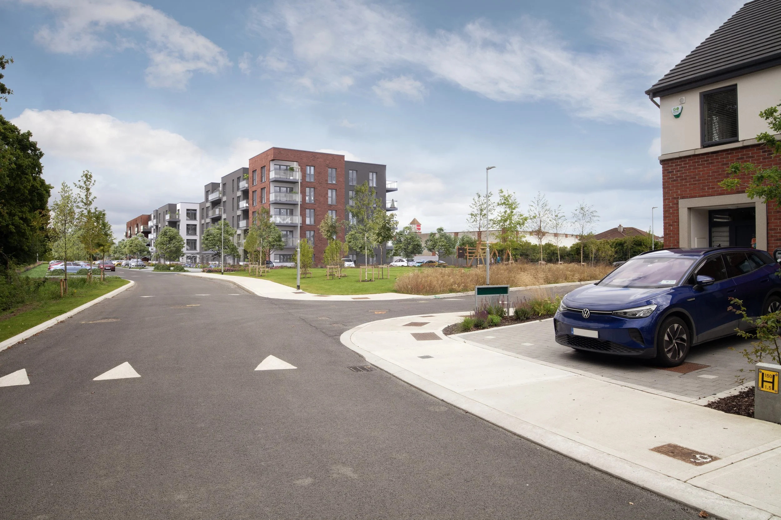 Residential neighborhood with modern apartment buildings, parked cars, green trees, and a playground in the background on a bright day.