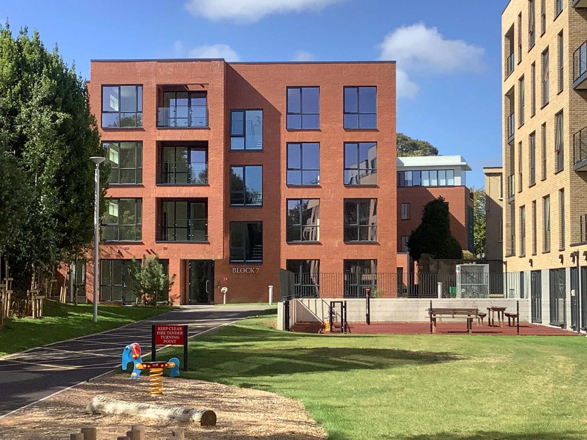 Modern red brick apartment building labeled 'Block 7' with surrounding green lawns, playground, and outdoor seating area, under a partly cloudy sky.